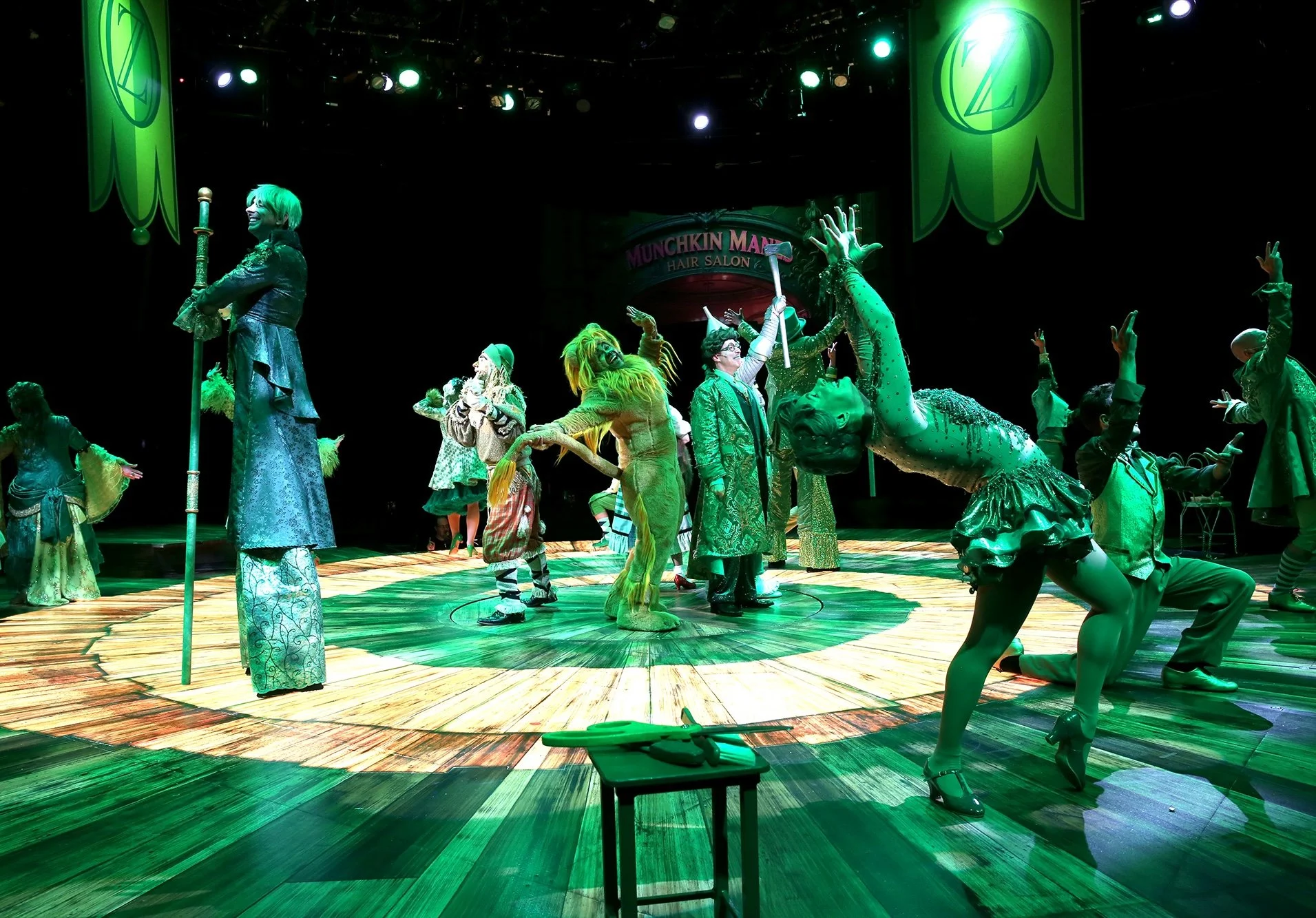 Performers dressed in colorful, whimsical costumes on a stage with a backdrop reading 'Munchkinland' in a theatrical production, with green lighting and a wooden floor.