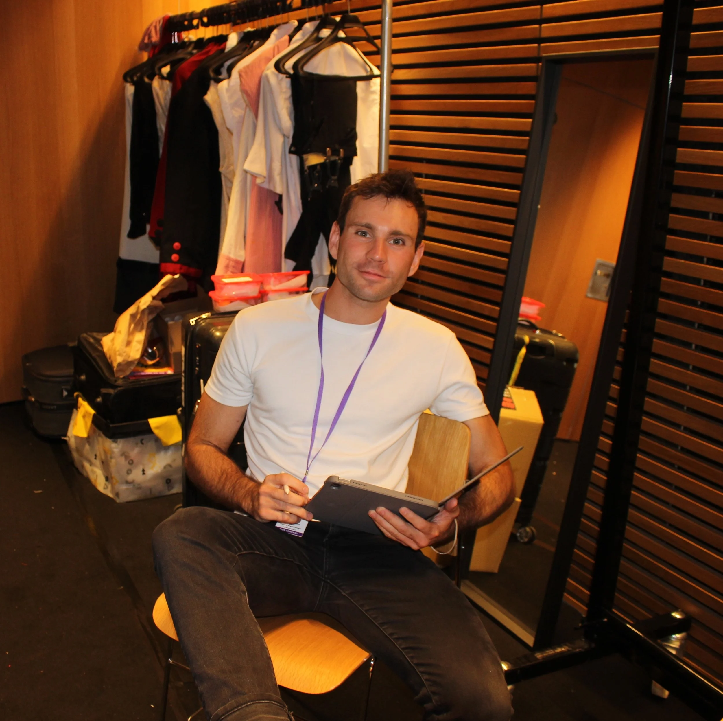 Man sitting on a yellow chair, holding a presentation remote, in front of a coat rack with clothes hanging, in a room with wooden wall paneling.
