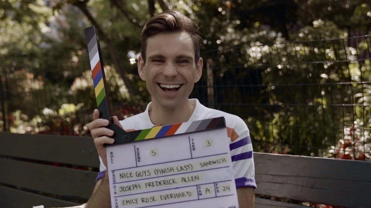 A young man sitting on a park bench holding a clapperboard and smiling at the camera.