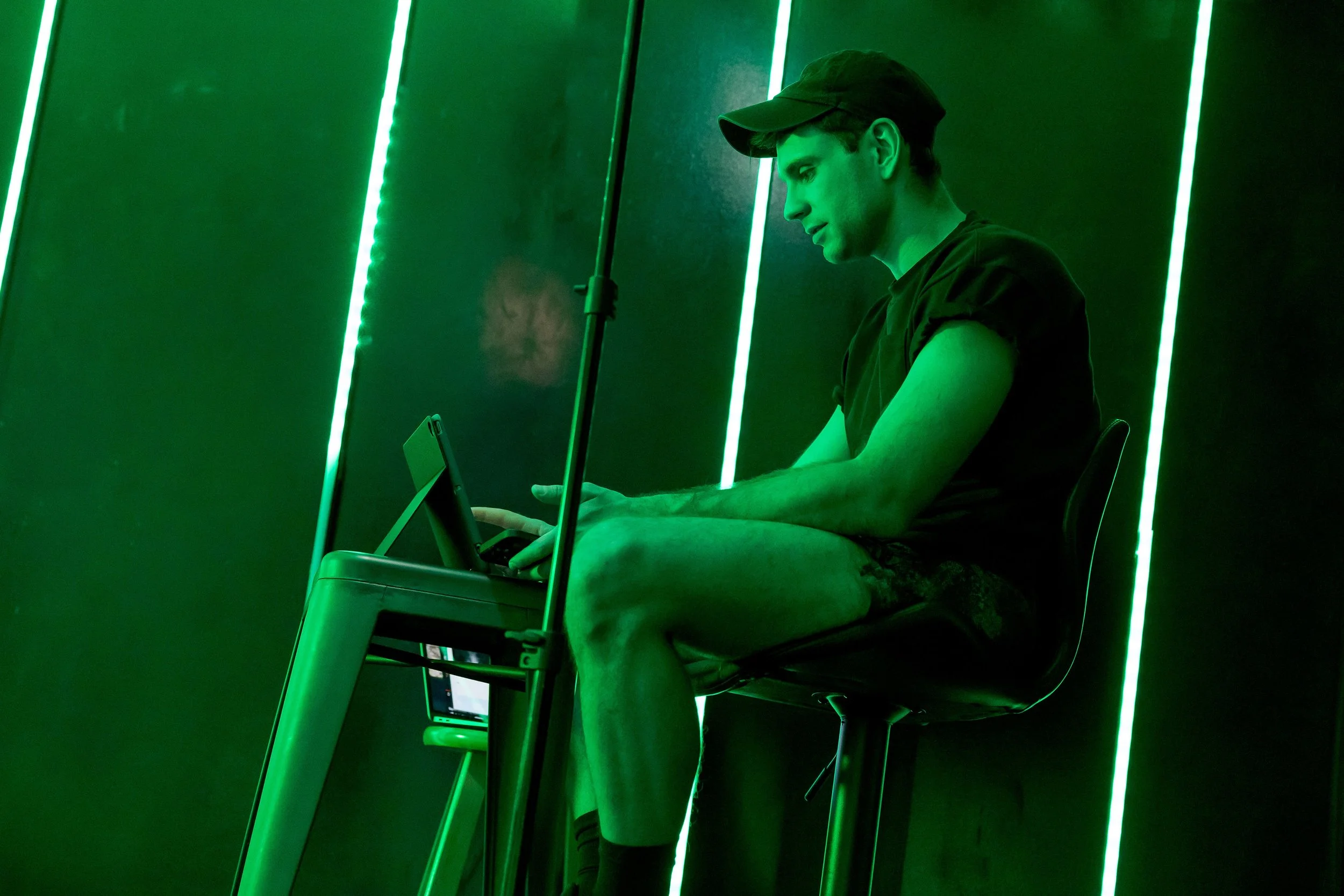 A young man sitting on a chair, wearing a black cap and black T-shirt, looking at his phone with a focused expression, in a room illuminated by green LED lights.