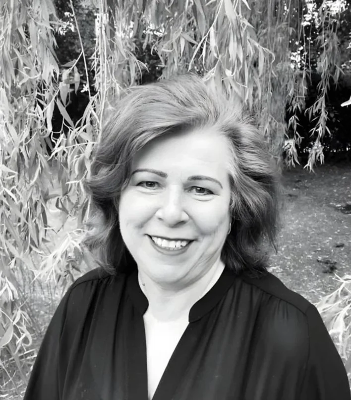 A woman with shoulder-length hair smiling outdoors, with trees and foliage in the background.
