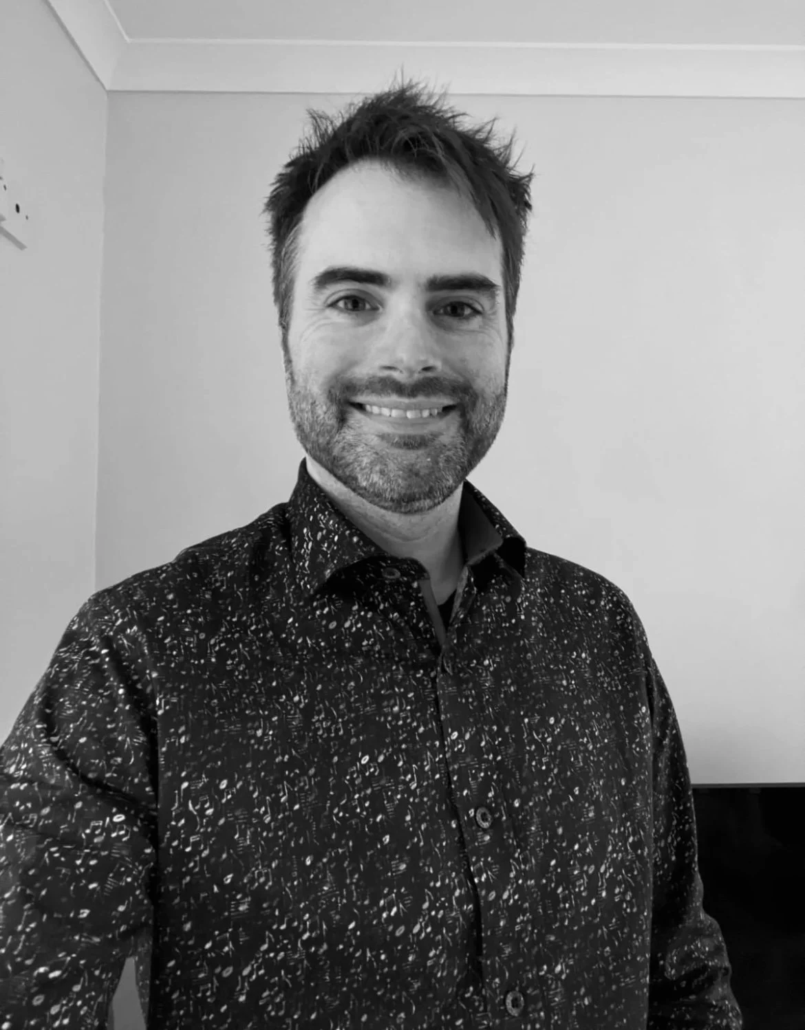 Black and white photo of a man with dark, messy hair and a beard, smiling, wearing a patterned button-up shirt, standing in a room with plain walls.