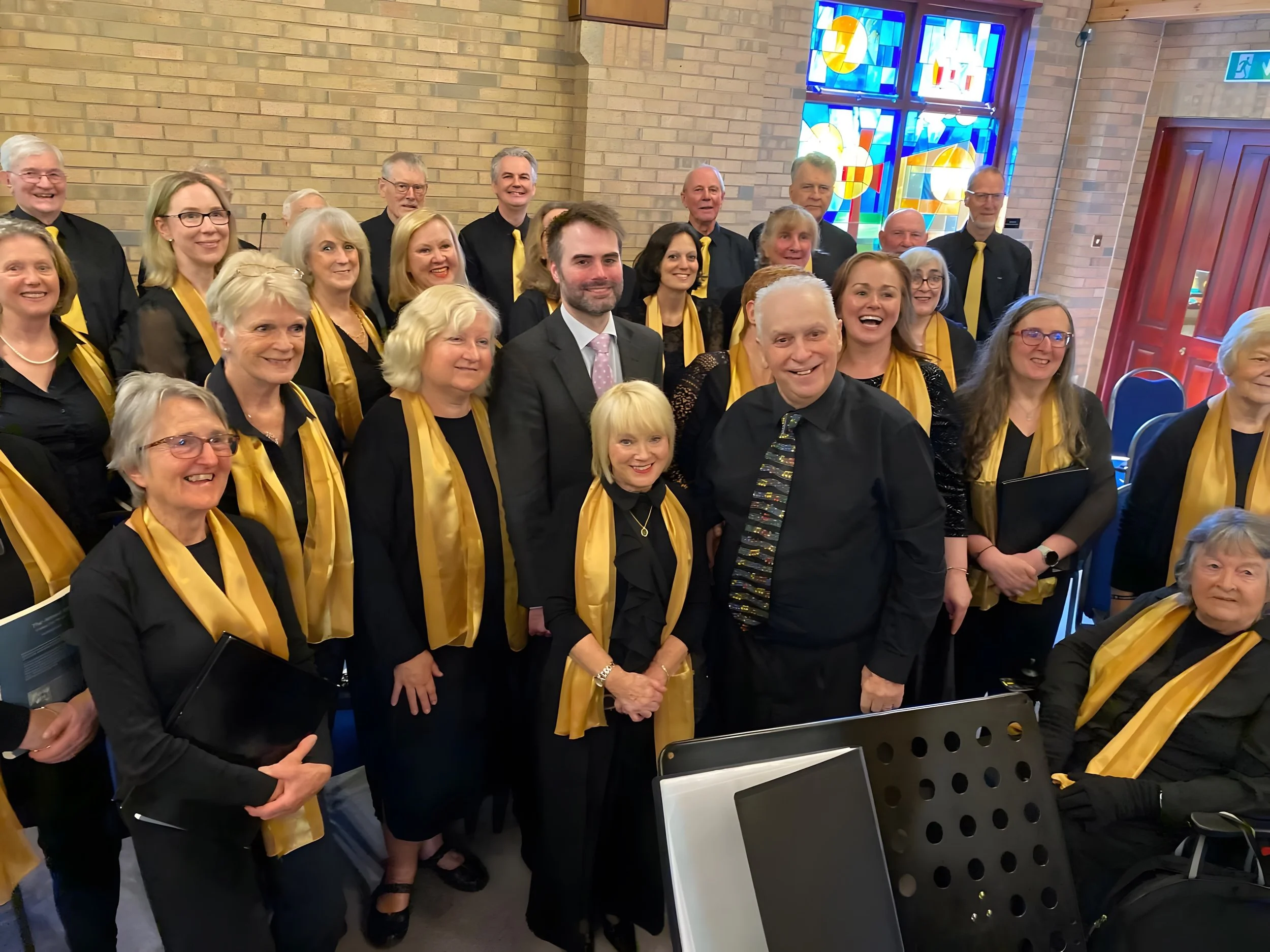 A group of cheerful choir members dressed in black with yellow scarves, standing inside a church with brick walls and stained glass windows.
