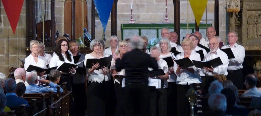 A choir of mainly older adults singing in a church, led by a conductor standing in front, with audience seated in pews watching.