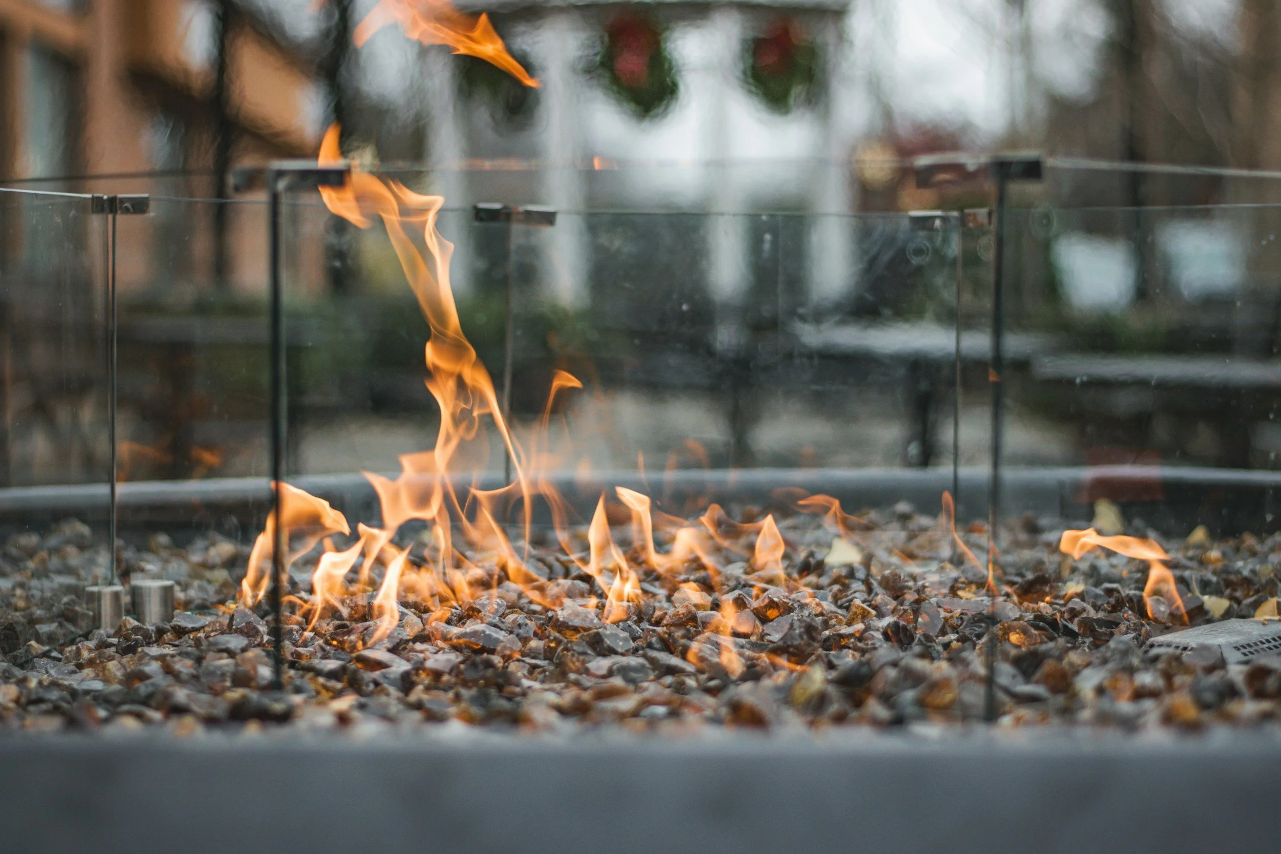 Close-up of a gas fireplace with visible flames, viewed through glass, with a gravel base and blurred outdoor background. Outdoor firepit. outdoor fireplace.