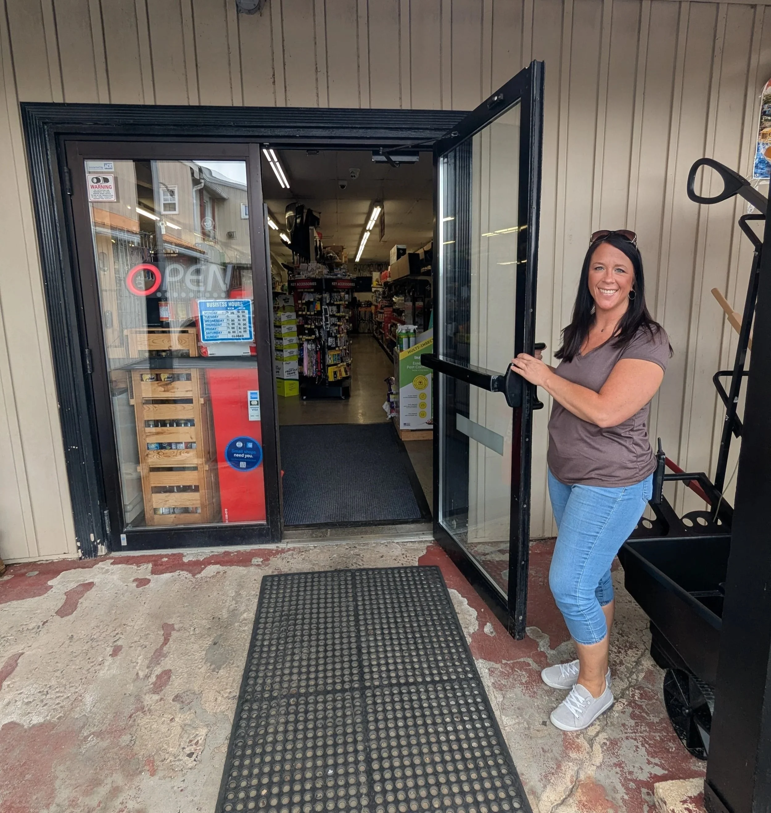 A woman is standing outside a store with a glass door, holding the door open. She is smiling, wearing a gray t-shirt, blue jeans, and white sneakers. The store's interior is visible through the door, showing product shelves, signs, and store hours on the door.