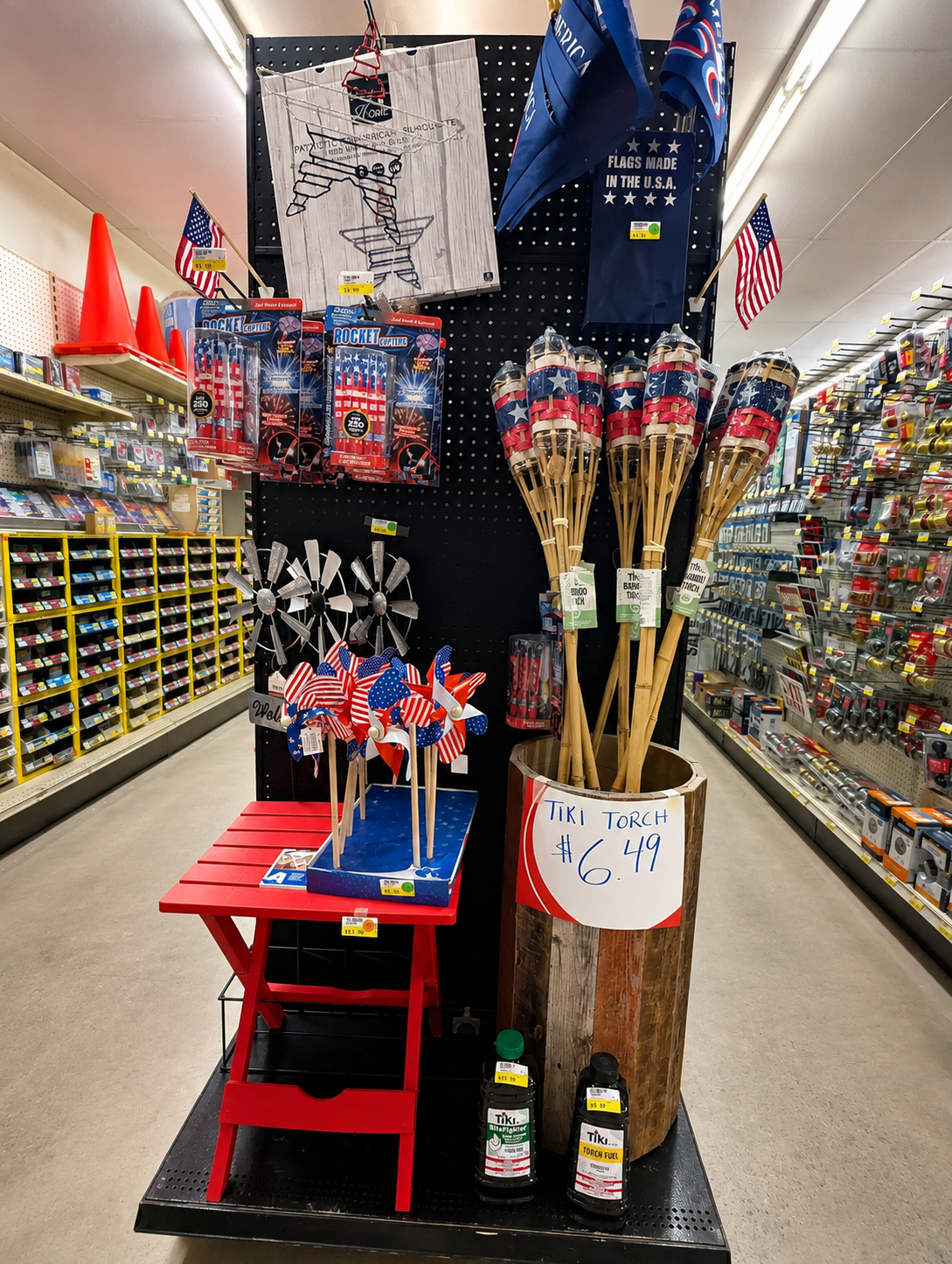 Red wooden table displaying colorful pinwheels with American flag designs, set in a hardware store aisle with various tools and flags. Seasonal