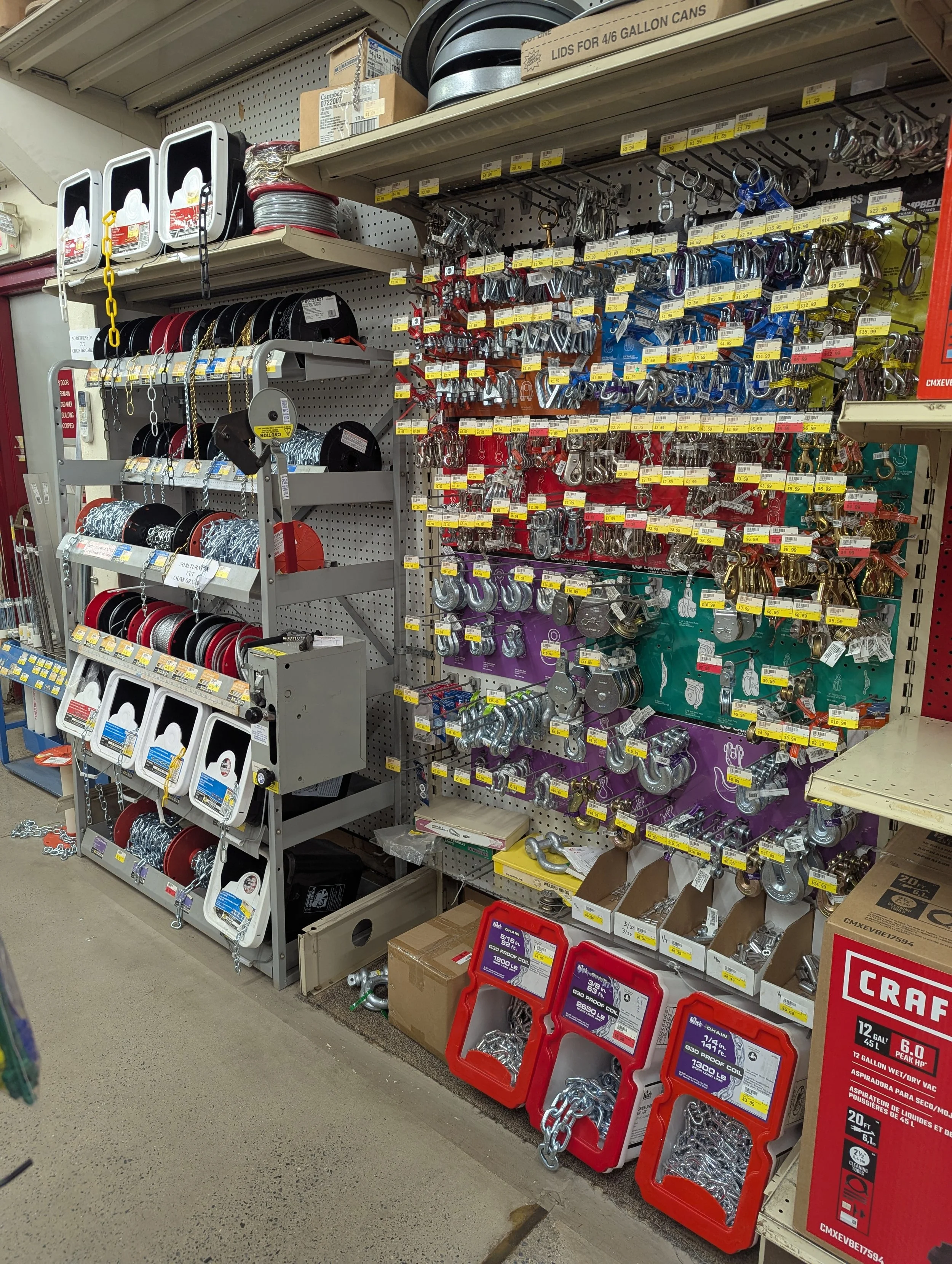 A store aisle displaying various mechanical hooks, chains, and fittings on shelves and in bins used for industrial and hardware purposes.