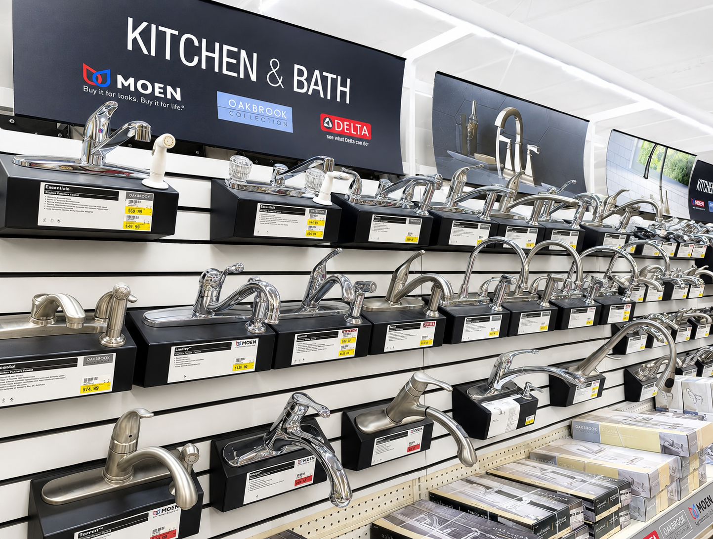 Display of various kitchen faucets on a slatwall at a retail store. A large sign above reads 'KITCHEN & BATH' with brand logos for Moen, Oakbrook Collection, and Delta.