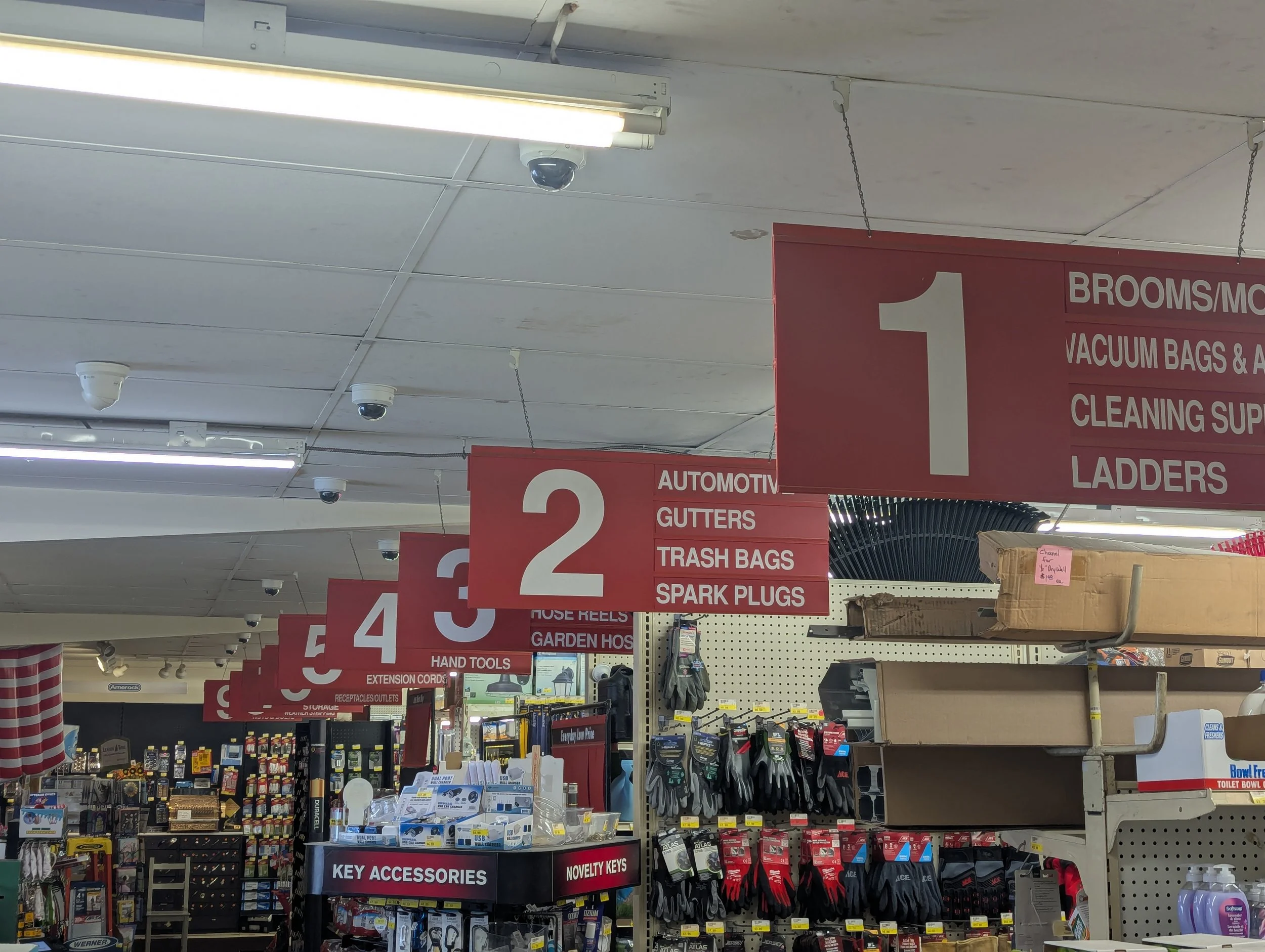 Inside an auto parts store, showing aisle signs for categories such as Brooms, Vacuum Bags, Cleaning Supplies, Ladders, and Automotive Gutter, Trash Bags, Spark Plugs, and Key Accessories.