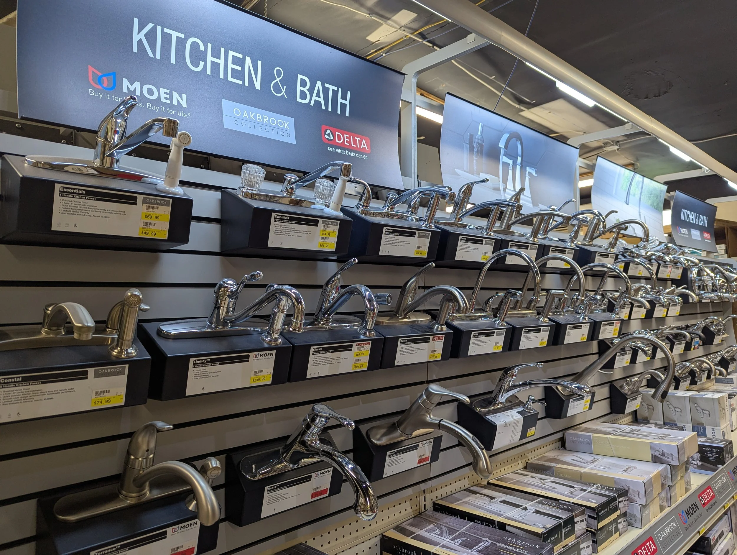 Display of kitchen and bathroom faucets arranged on shelves in a store, with signage indicating brands Moen and Delta.