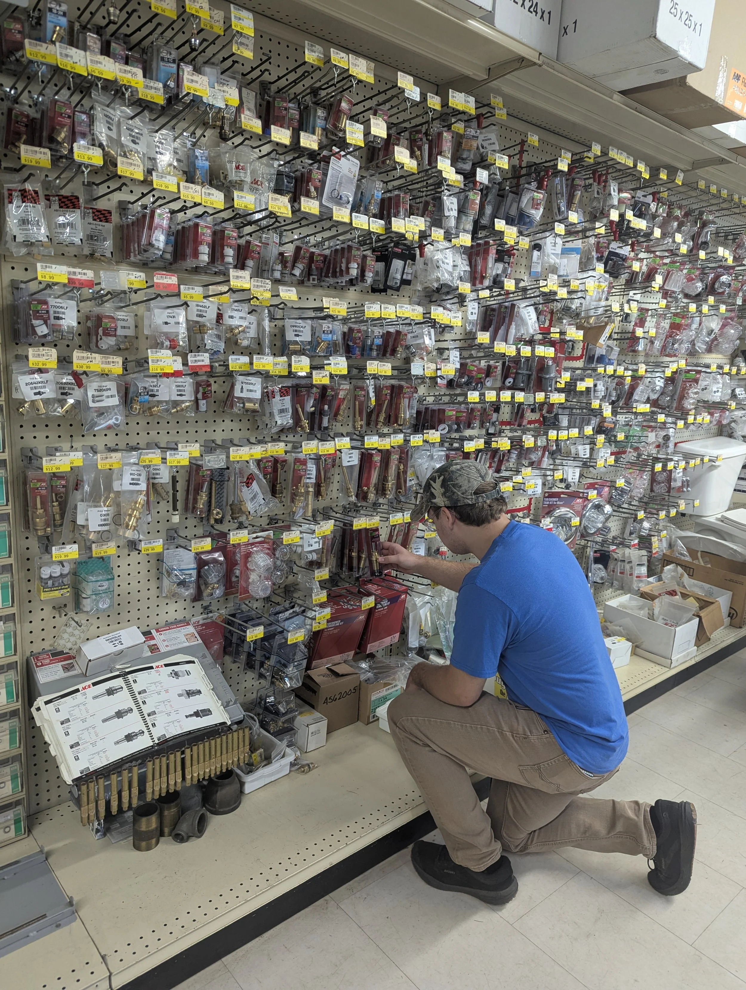 A man wearing a blue shirt, beige pants, and a camouflage baseball cap is kneeling in front of a display of plumbing parts and fittings at a hardware store. The display includes various valves, connectors, and pipes, with prices tagged on yellow labels. The man appears to be examining or selecting an item from the shelf.