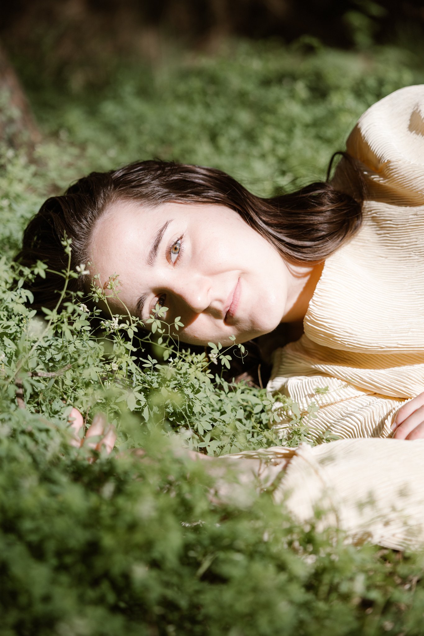 Close-up van een vrouw liggend op het gras, met glimlach en omringd door groene planten.