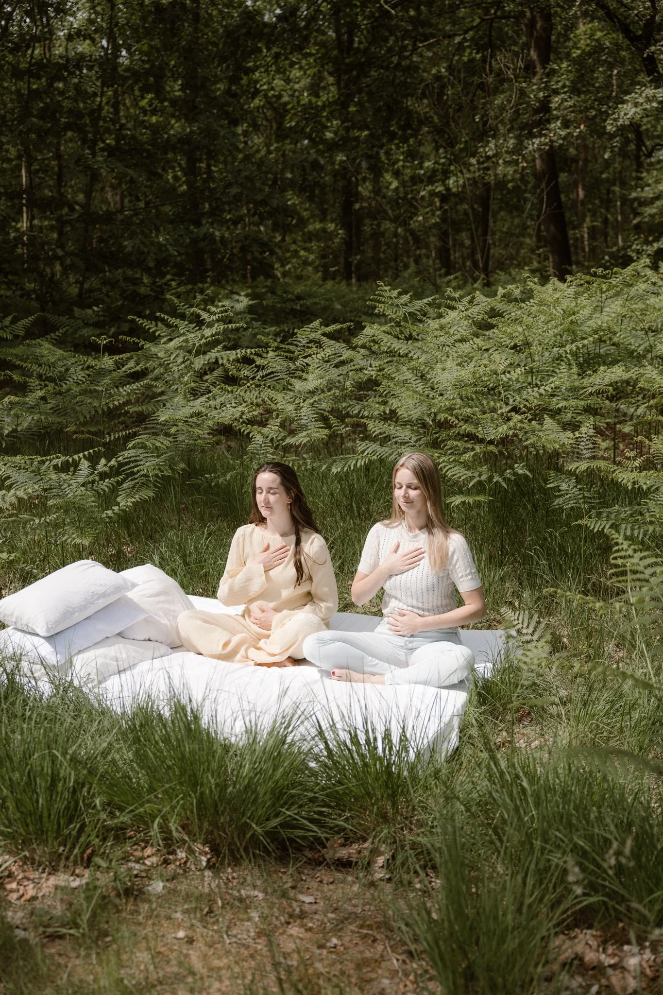 Twee jonge vrouwen zitten op een witte mat op gras in een bos, mediteren met handen op de borst, omringd door groene planten en bomen.