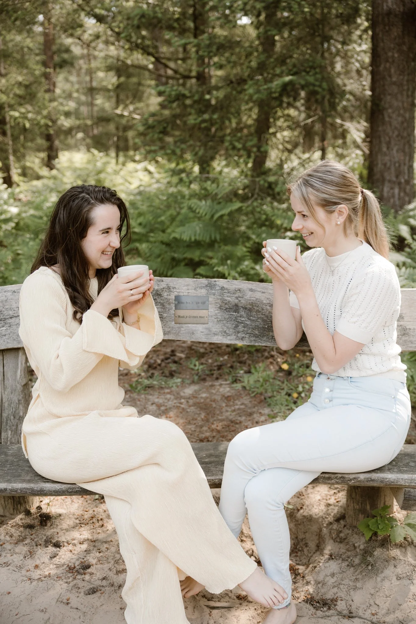 Twee jonge vrouwen zitten op een bankje in het bos, lachen en drinken uit witte bekers.