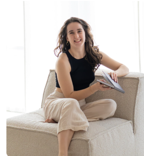 Vrouw zittend op een beige bank, lachend, met een boek in haar handen, in een lichte kamer met witte gordijnen.