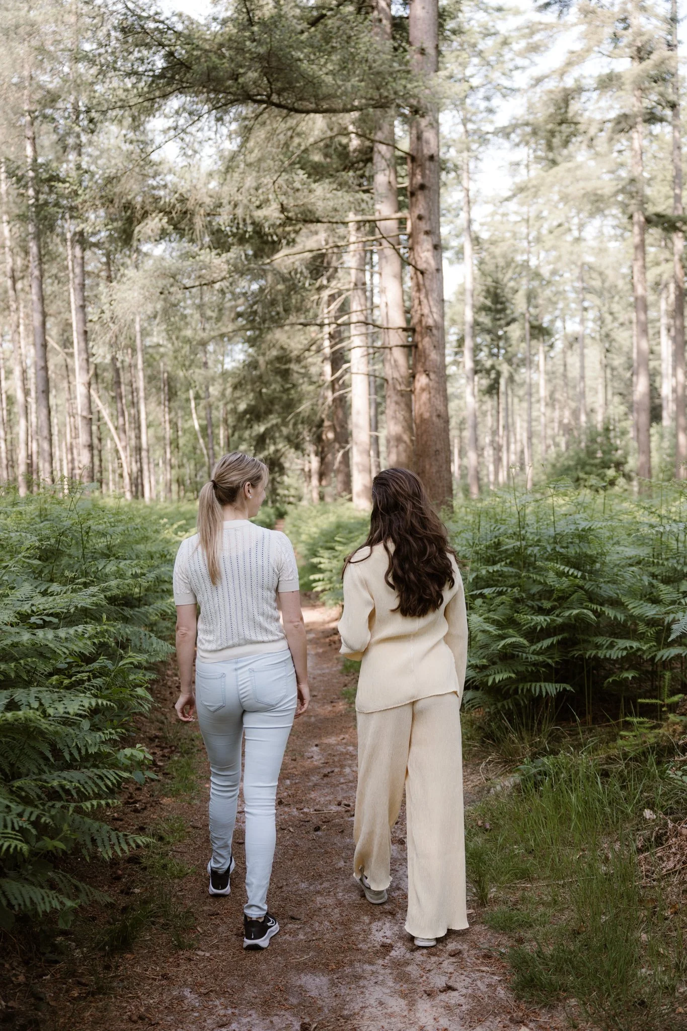 Twee vrouwen wandelen in een bos, gezien van achter, met groene planten en bomen om hen heen.