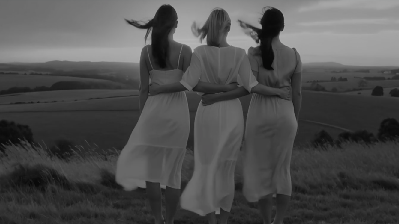 Drie vrouwen in witte jurken lopen arm in arm over een veld met een landschap op de achtergrond, allemaal met de haren die in de wind waaien, zwart-wit foto.