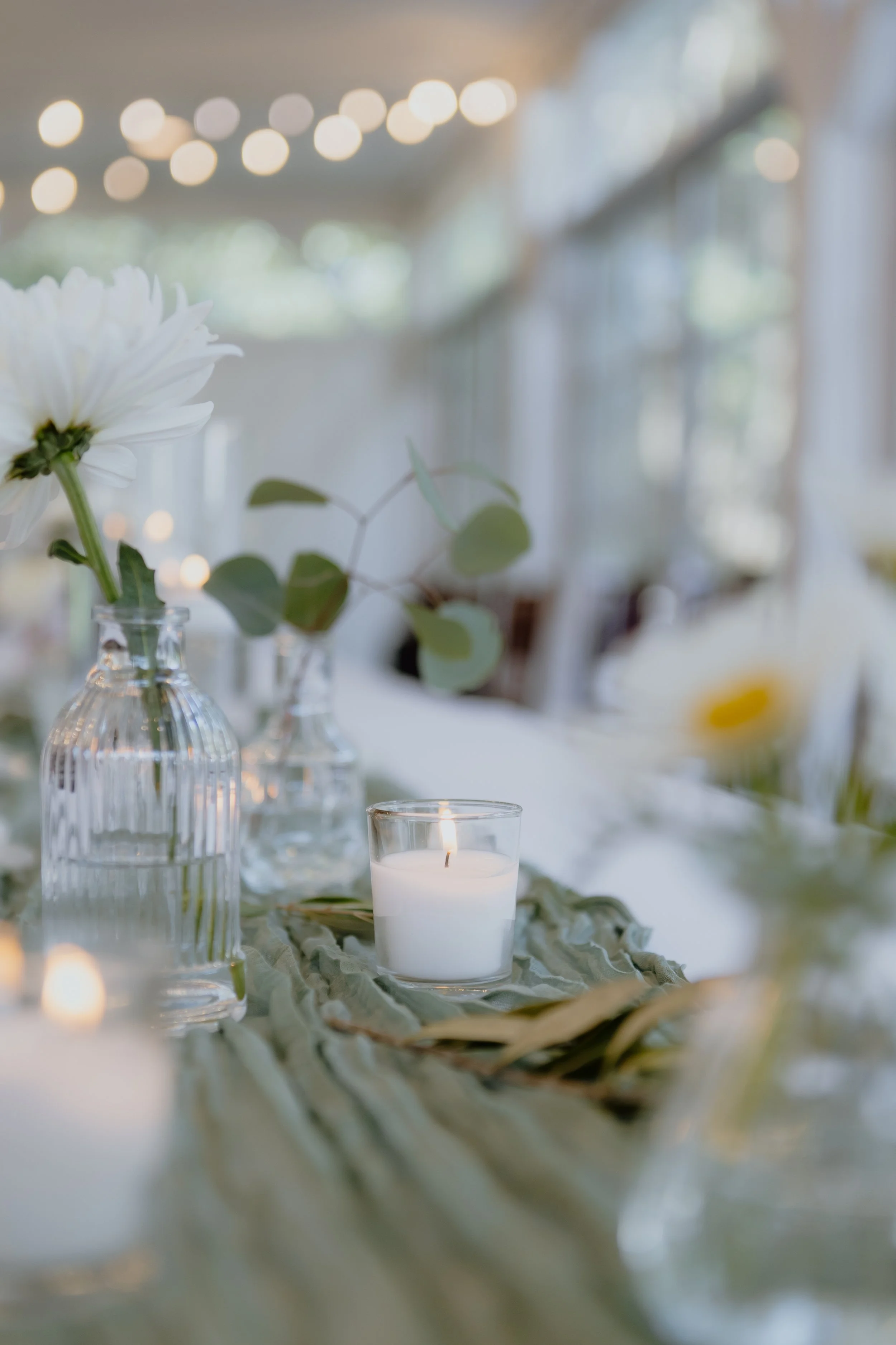 Close-up of a white flower in a glass vase on a table at a softly lit event or gathering.