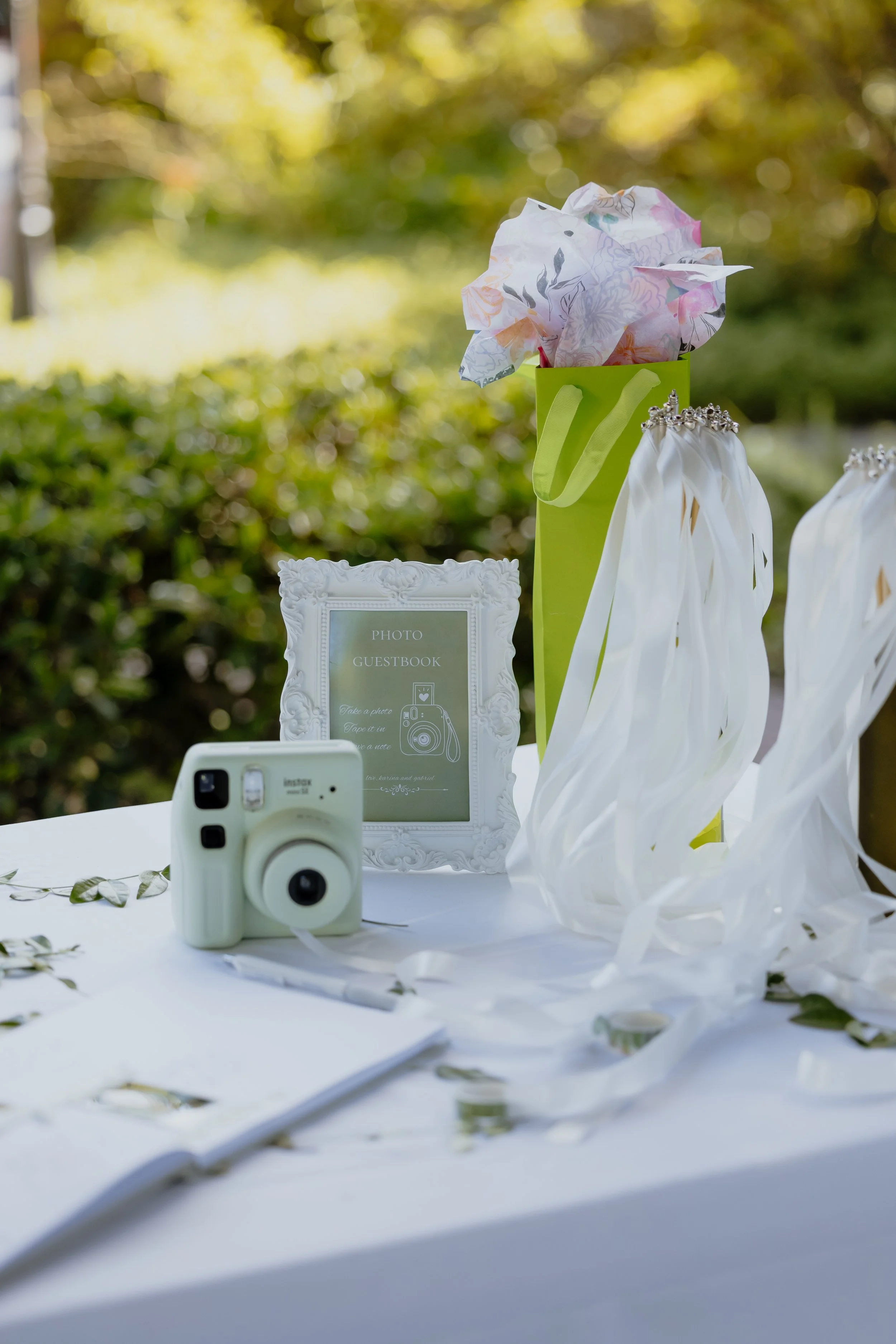 Decorative wedding table setup with a white photo guestbook, a green camera, a green gift bag with crumpled tissue paper, a white gift bag with ribbon handles, and scattered leaves.