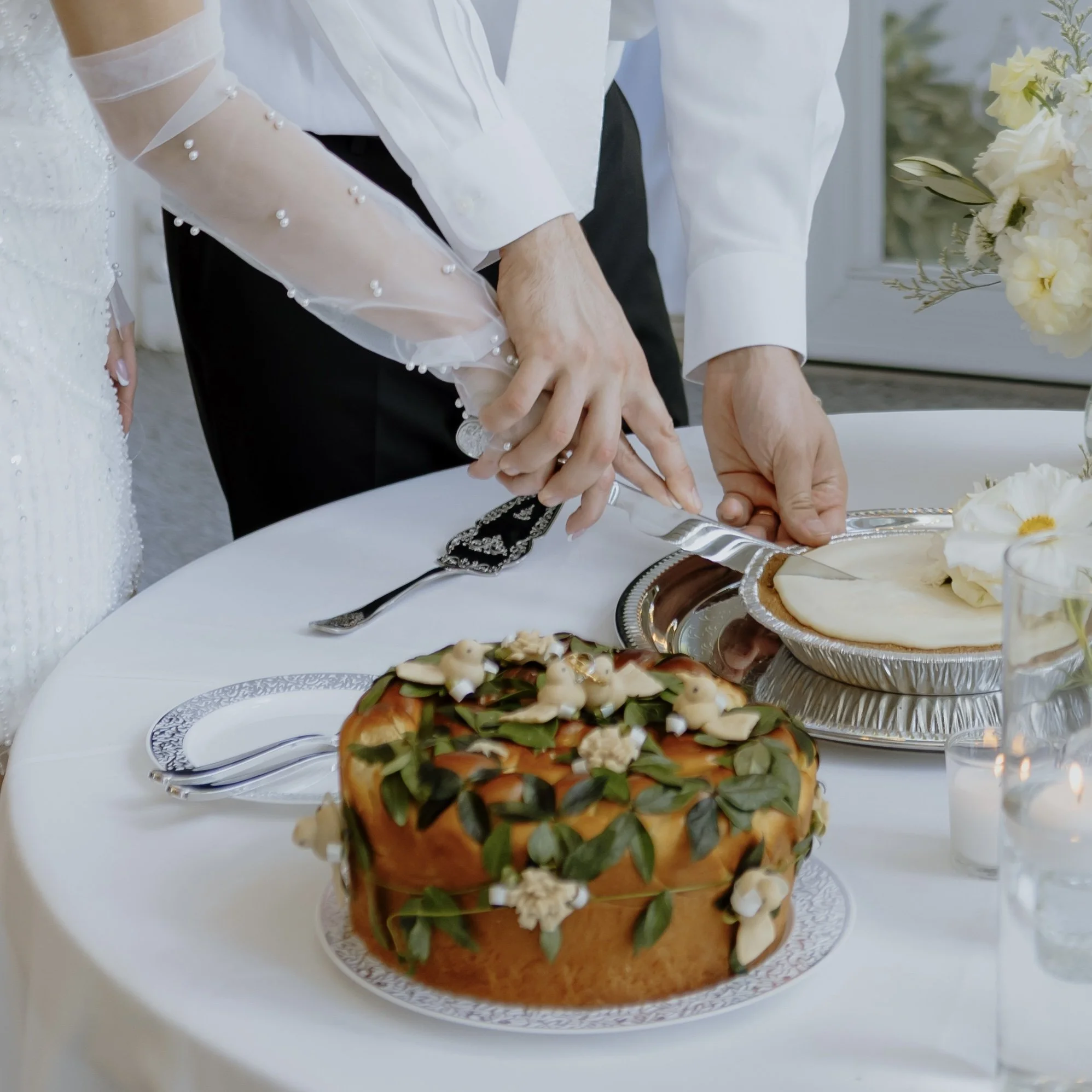 Close-up of a korovai, decorated with green leaves and beige icing, and a bouquet of white flowers in the background. The korovai is on a decorated silver tray on a white table.