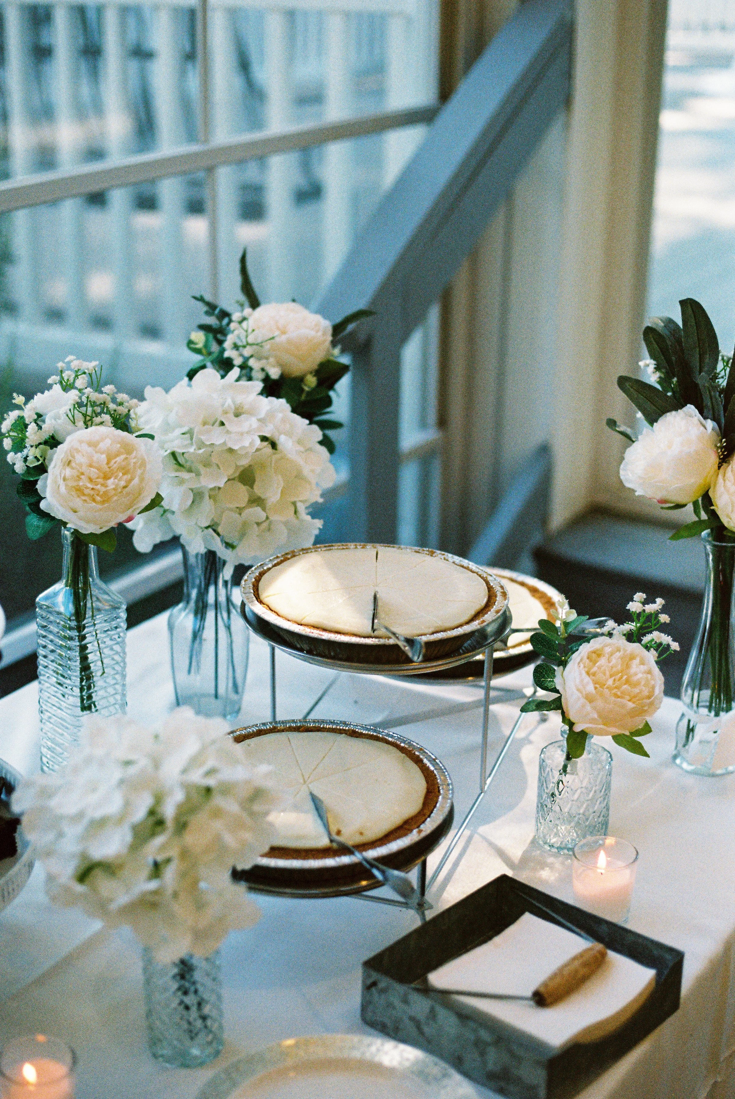 A table decorated with white floral arrangements, candles, and wooden pies in a bright room with large windows.