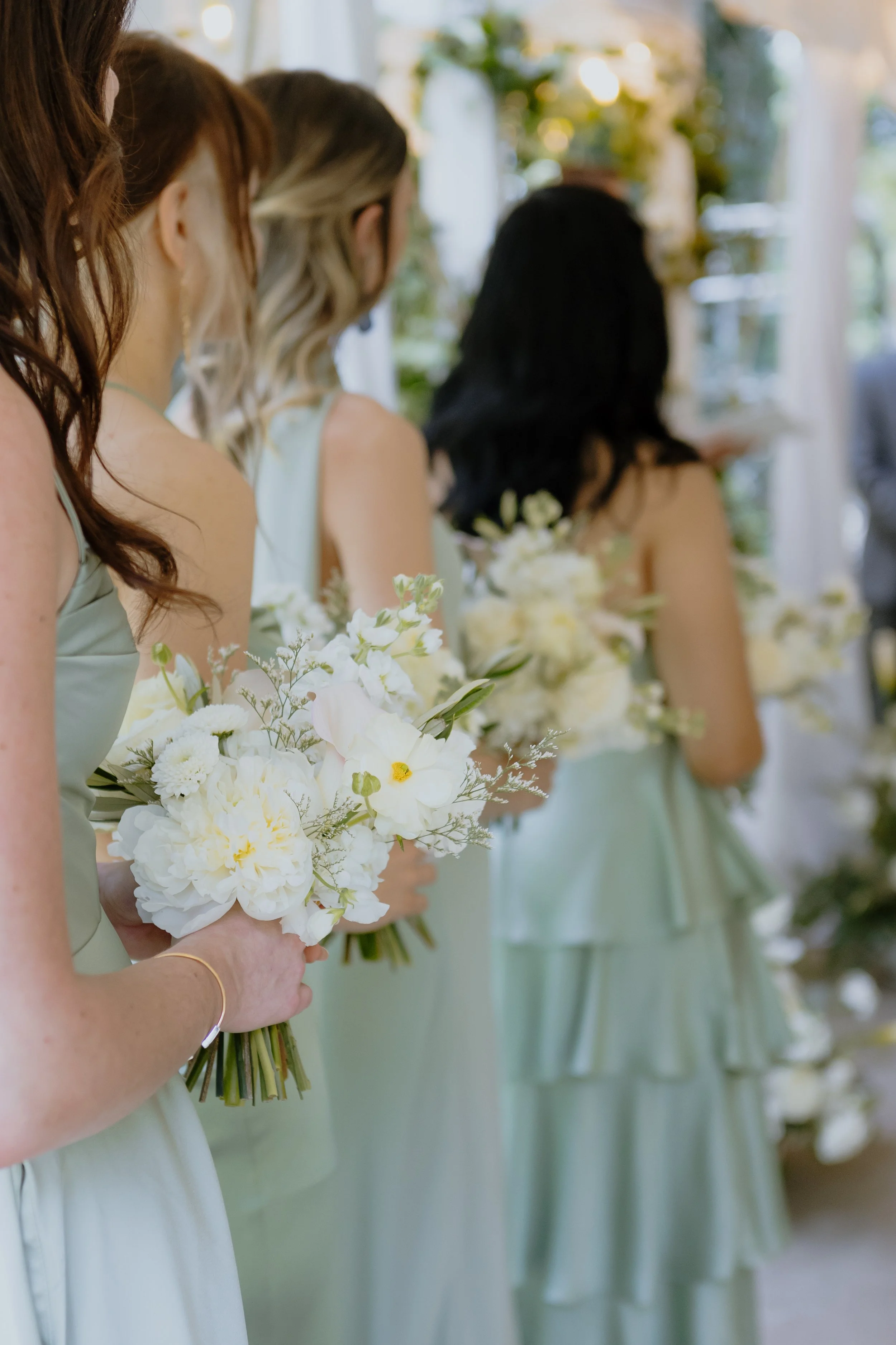 Four women in sage green dresses holding bouquets of white flowers, standing in a row at a wedding or formal event, indoors with natural light.