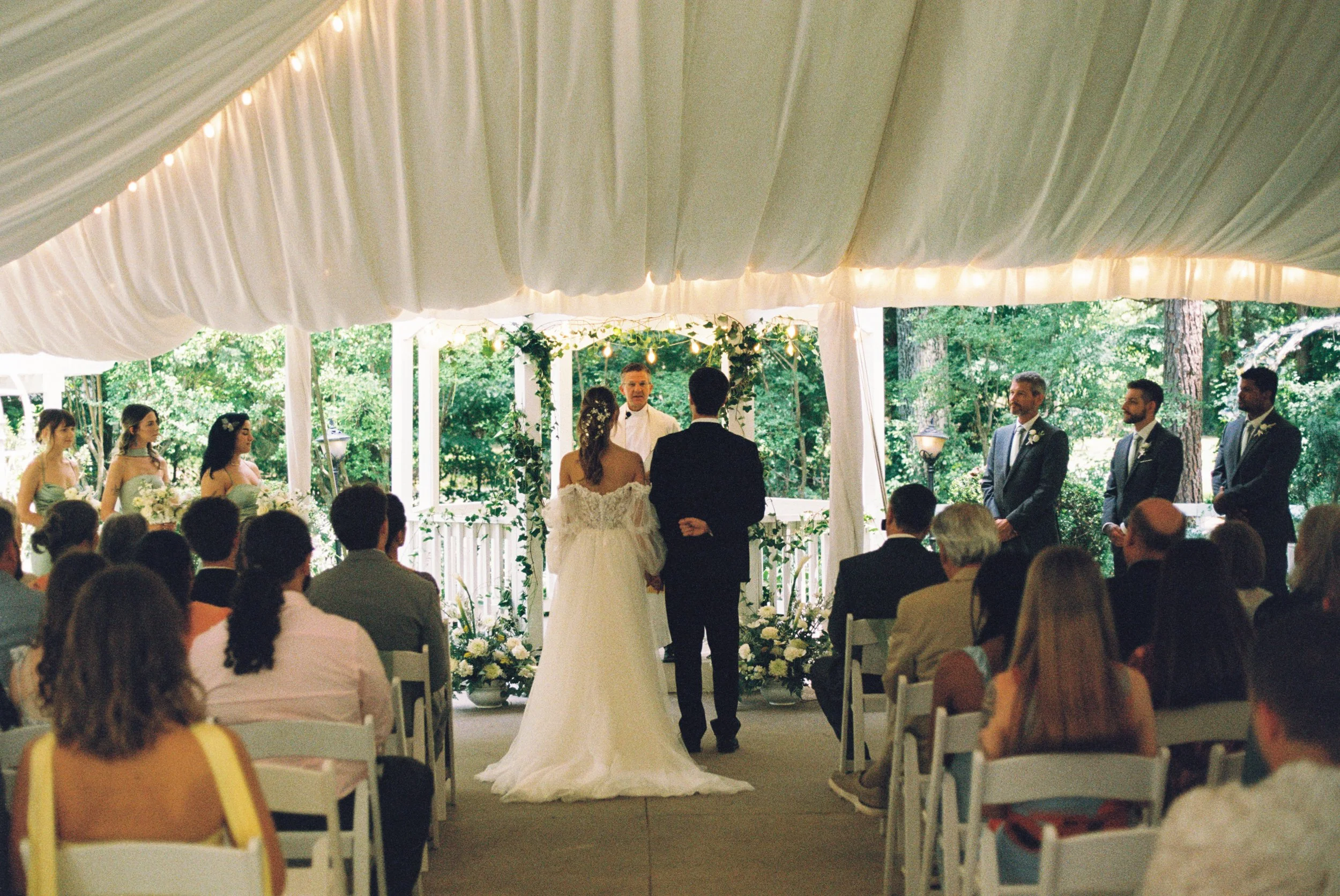 A wedding ceremony taking place under a white canopy with fairy lights, with the bride and groom standing before an officiant, surrounded by guests seated in white chairs in an outdoor setting with trees and greenery in the background.