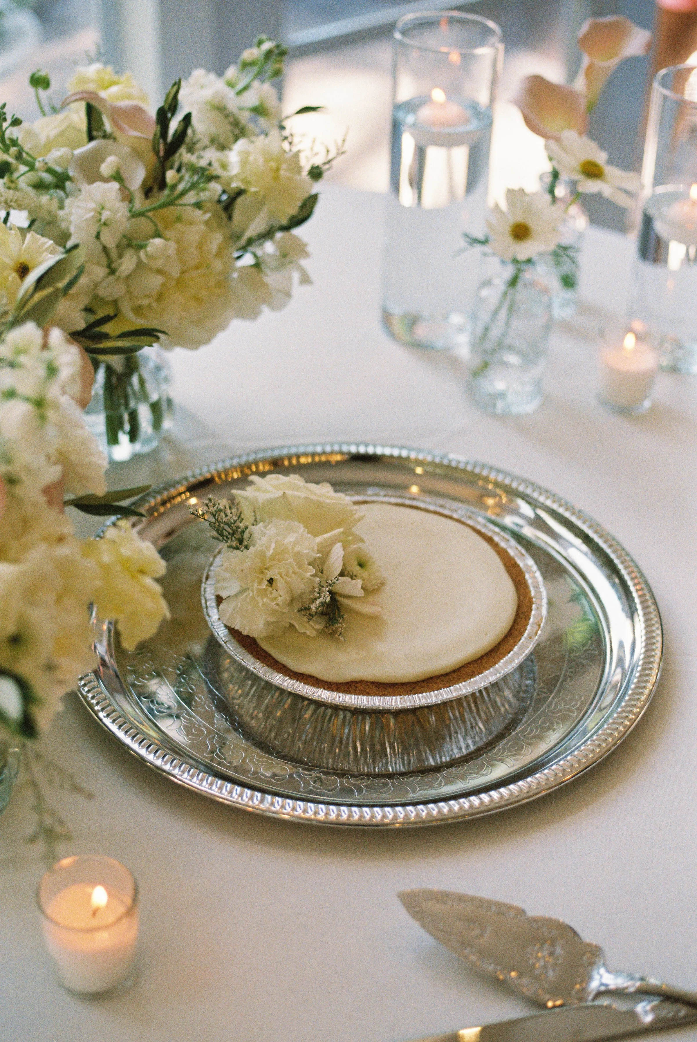 A table setting featuring a slice of a pie on a decorative silver plate, surrounded by white flowers in vases and small lit candles.