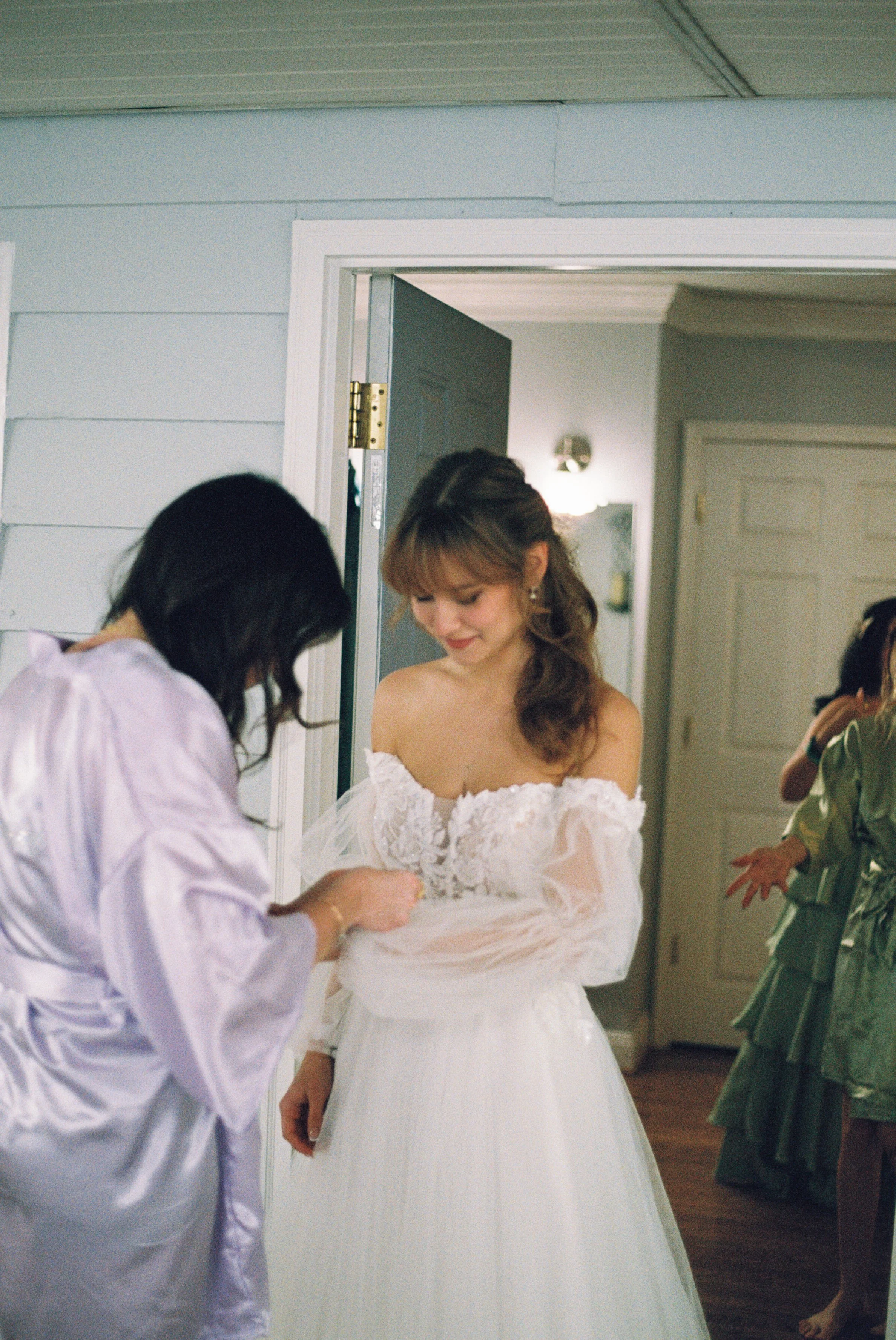 A woman in a white wedding dress is being helped by another woman with dark hair in a lavender robe. There are other people in the background in a room with light-colored walls and a partially open door.