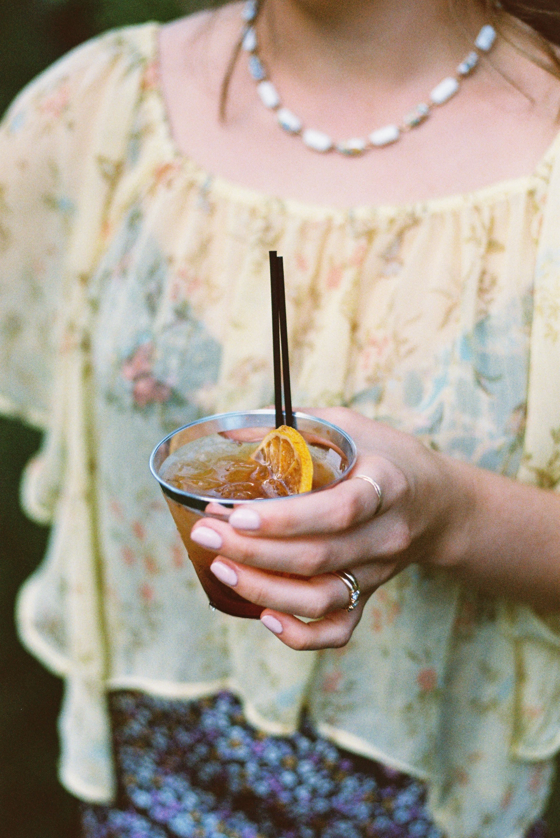 A woman wearing a yellow floral top holding a drink garnished with an orange slice and a straw.