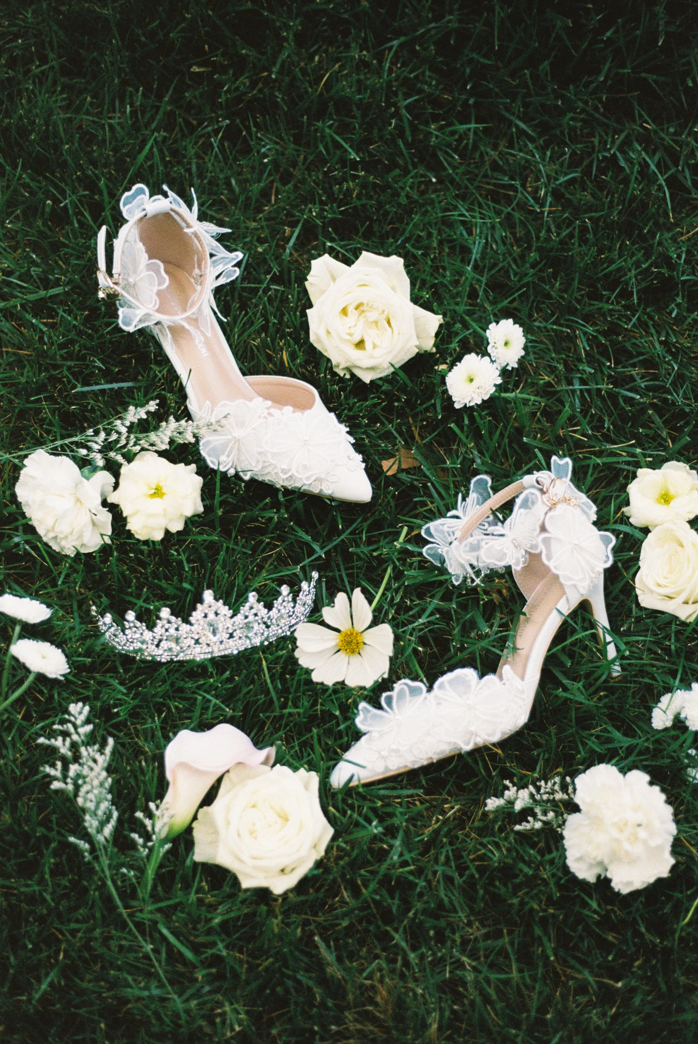 A white floral wedding shoe, a tiara, and white flowers placed on green grass.