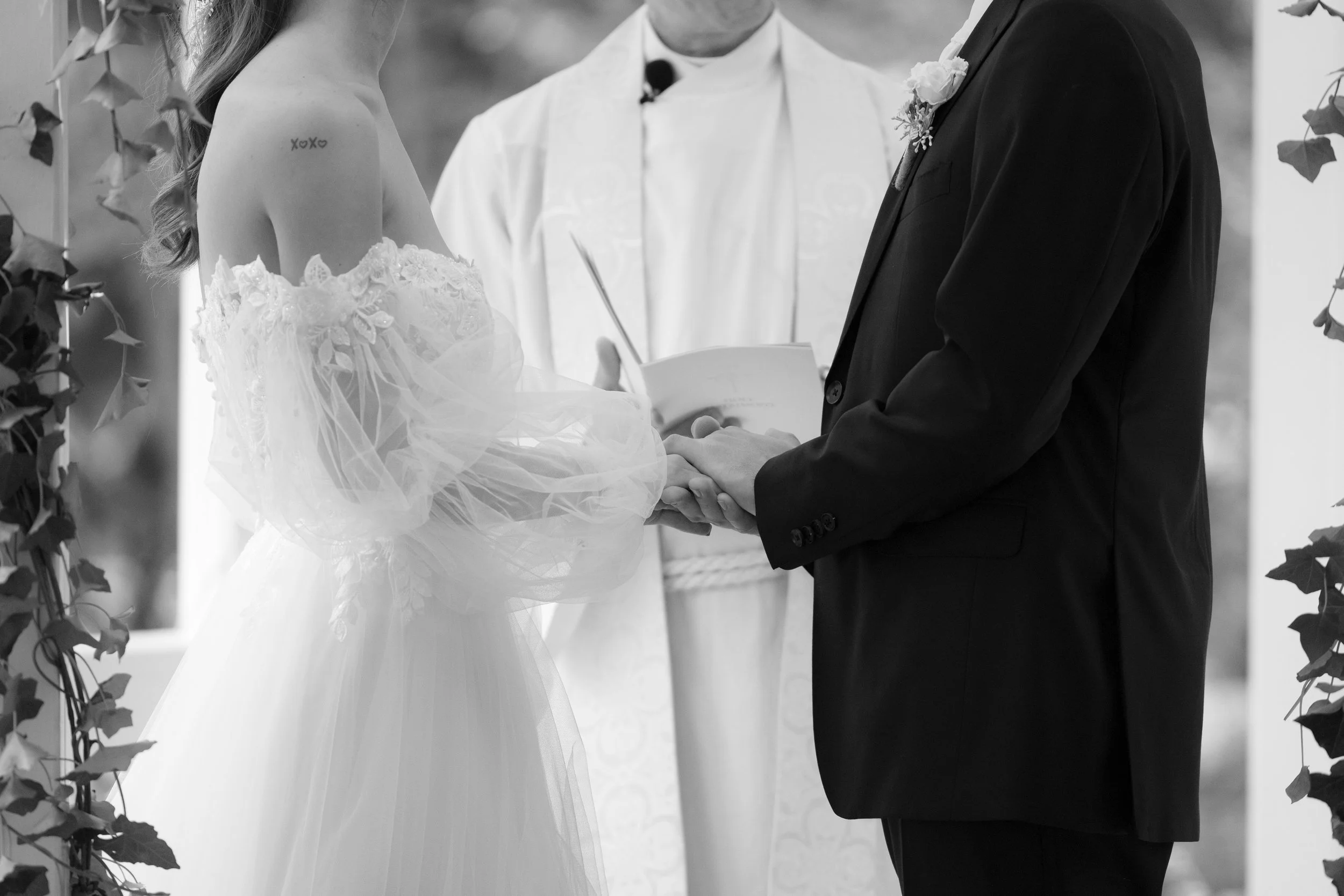A couple getting married, holding hands during the ceremony, with an officiant in the background. The bride is wearing a lace wedding dress with off-the-shoulder sleeves, and the groom is in a dark suit with a boutonniere. The picture is in black and
