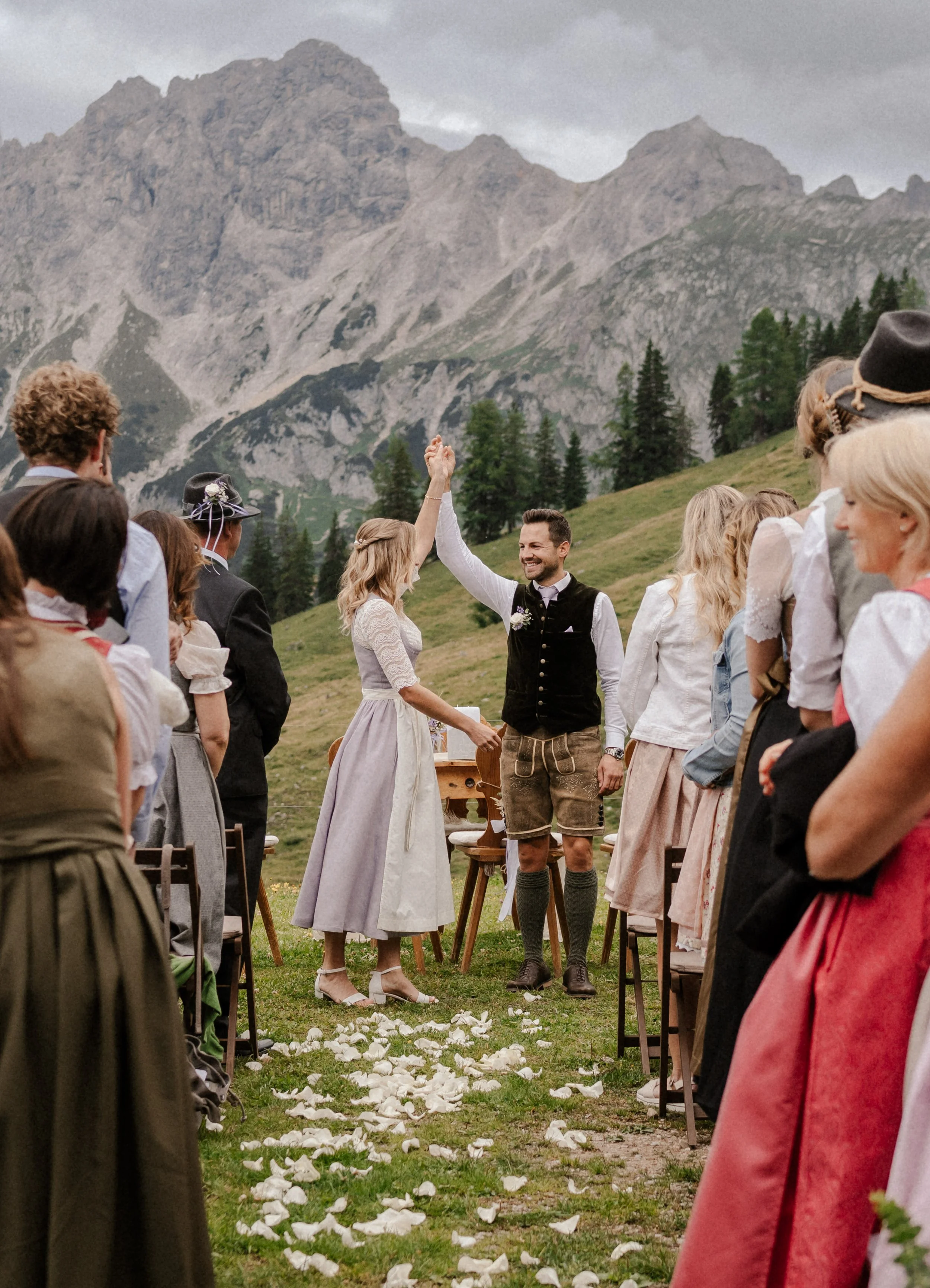 Ein Brautpaar bei einer Hochzeitszeremonie im Freien, umgeben von Gästen in traditioneller Kleidung, mit einer Bergkulisse im Hintergrund.