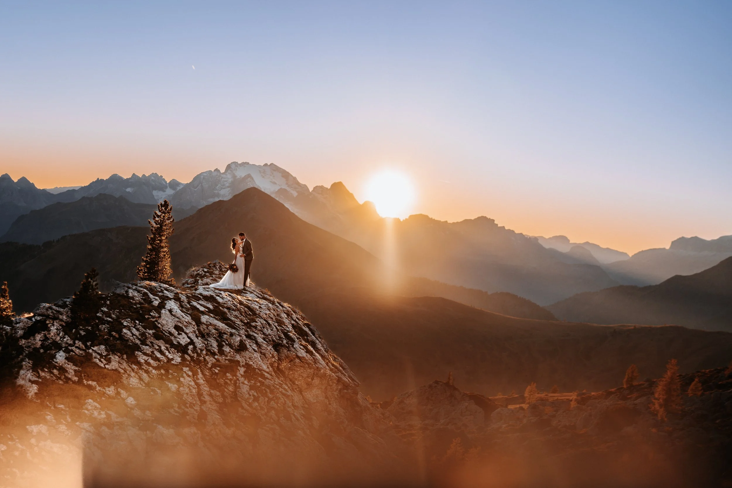 Ein Paar in Hochzeitskleidung steht auf einem Felsen vor einer Bergkulisse bei Sonnenuntergang.
