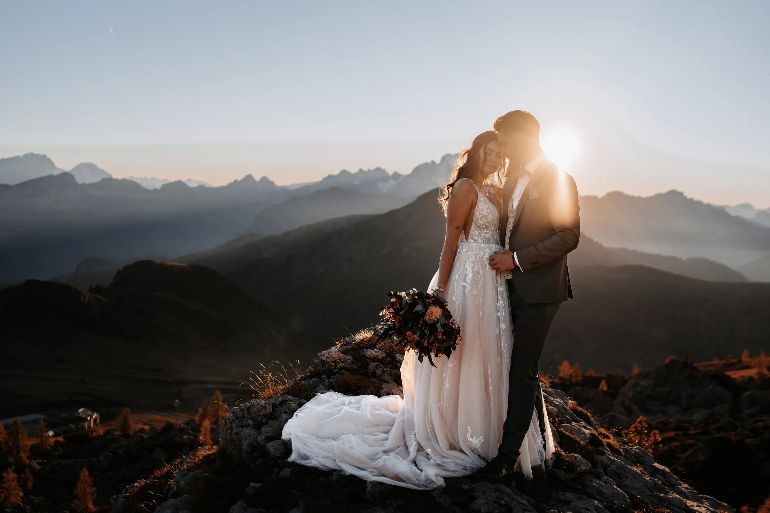 Ein Hochzeitspaar steht auf einem Felsen vor einer Berglandschaft bei Sonnenuntergang, sie umarmen sich und teilen einen intimen Moment.