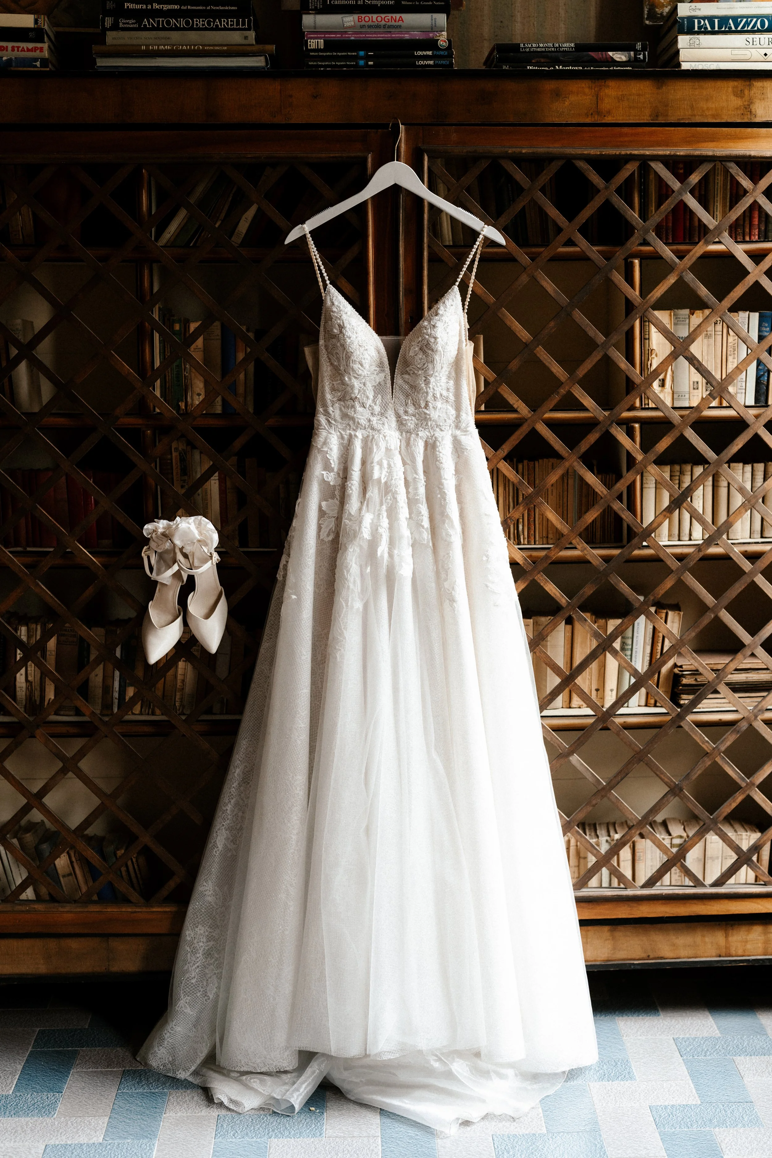 Ein weißes Brautkleid hängt an einem weißen Kleiderbügel vor einer Holzregalwand mit Büchern. Darunter hängen weiße Hochzeitsschuhe an einem Nagel.