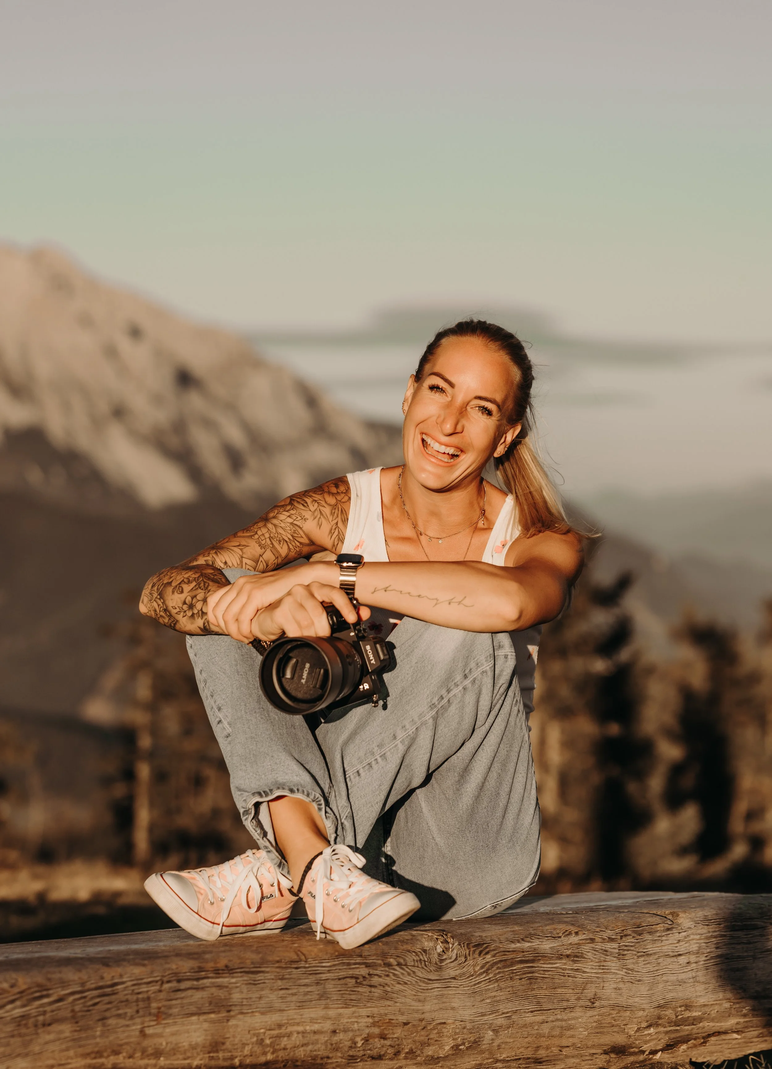Junge Frau mit Tattoos, sitzend auf einem Baumstamm, lachend und einen Fotoapparat haltend, im Freien bei Sonnenuntergang.