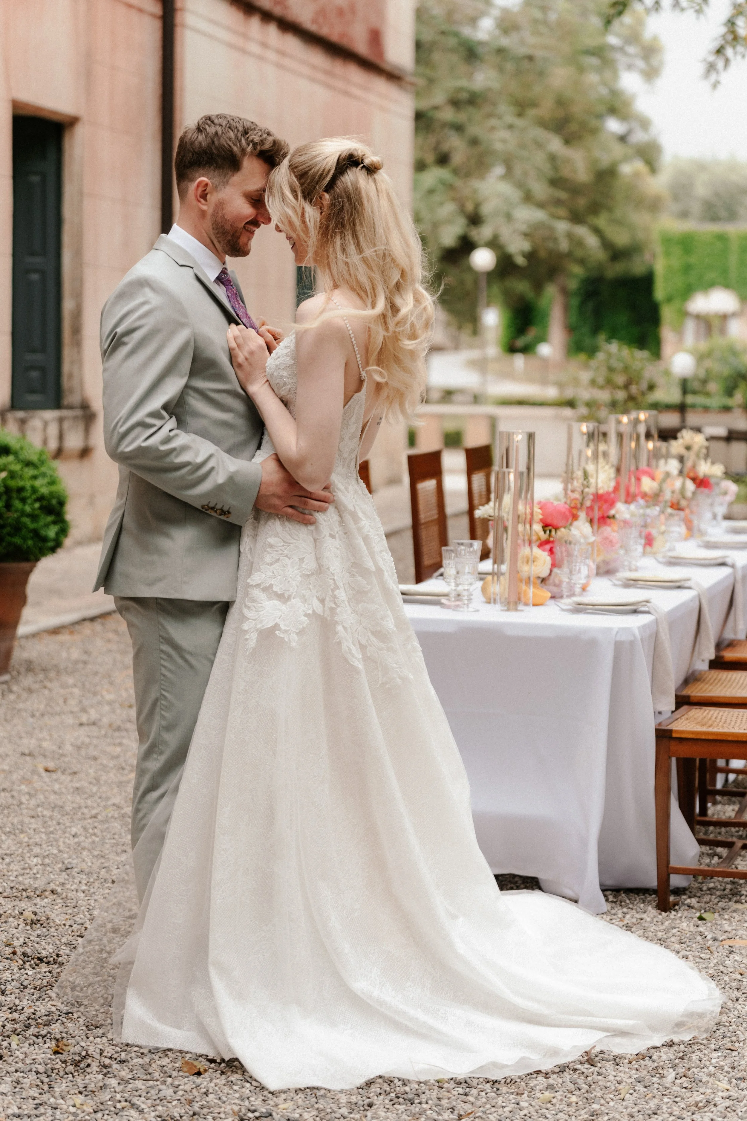Ein frisch verheiratetes Paar auf einer Hochzeit im Freien, das sich mit ihren Stirn aneinander lehnt, während sie lächeln. Im Hintergrund sind essgeschmückte Tische und grüne Bäume.