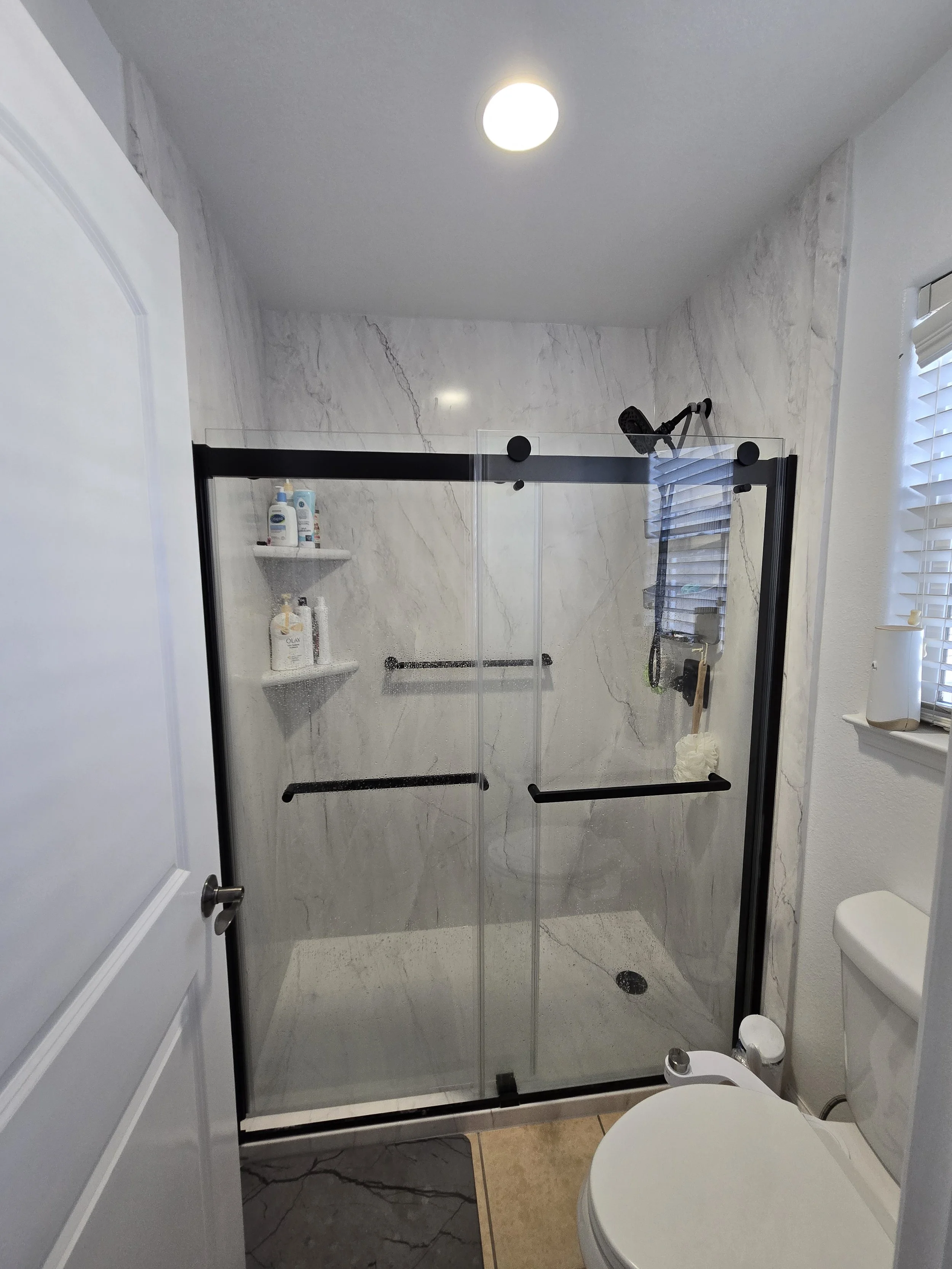 Small bathroom with a marble shower stall, a white toilet, and beige tiled floor. There are toiletries on a shelf inside the shower and a window with blinds on the right wall.