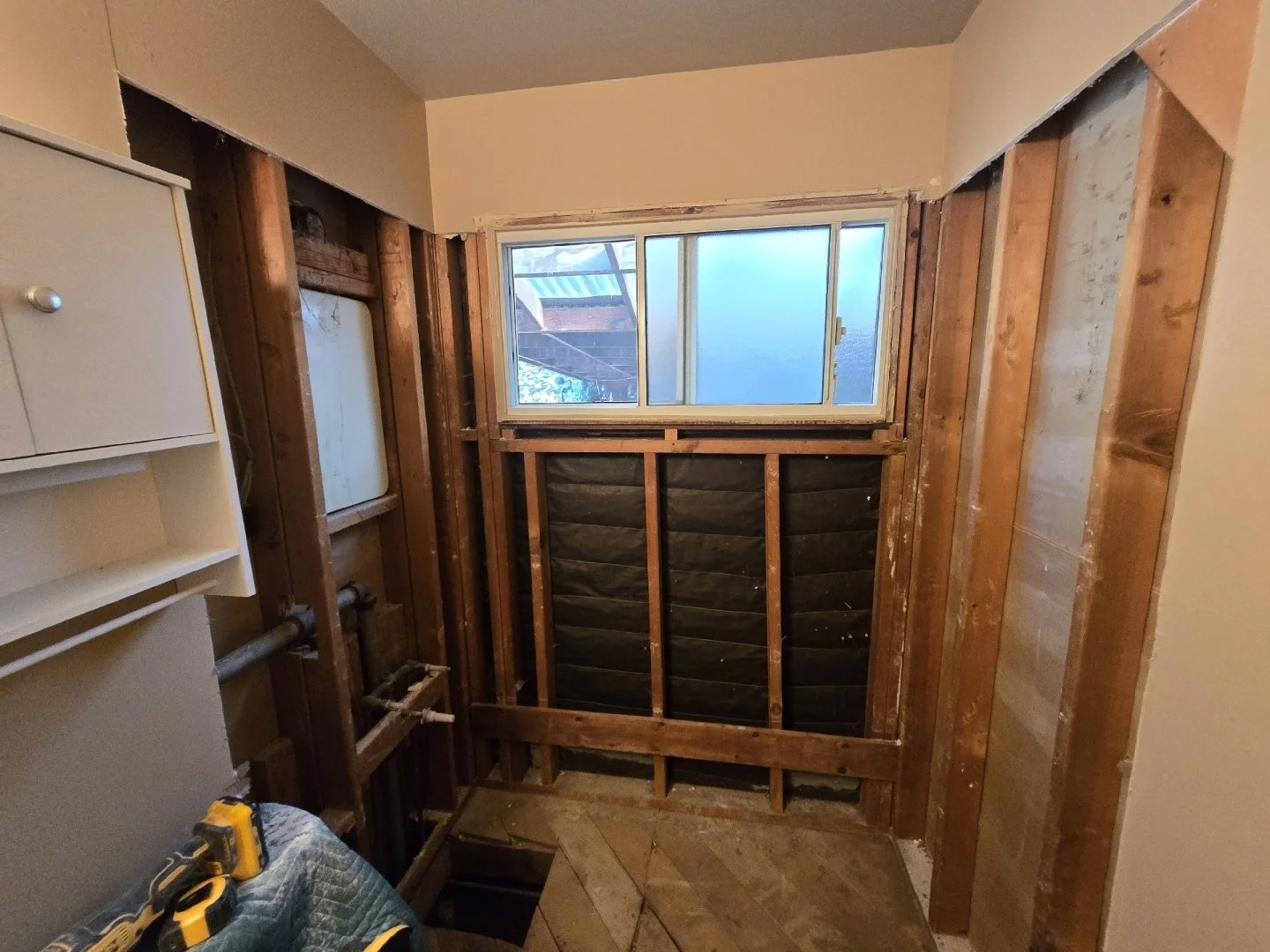 Room under renovation showing exposed wall studs, a window, and plumbing pipes, with construction tools on a quilted cushion.