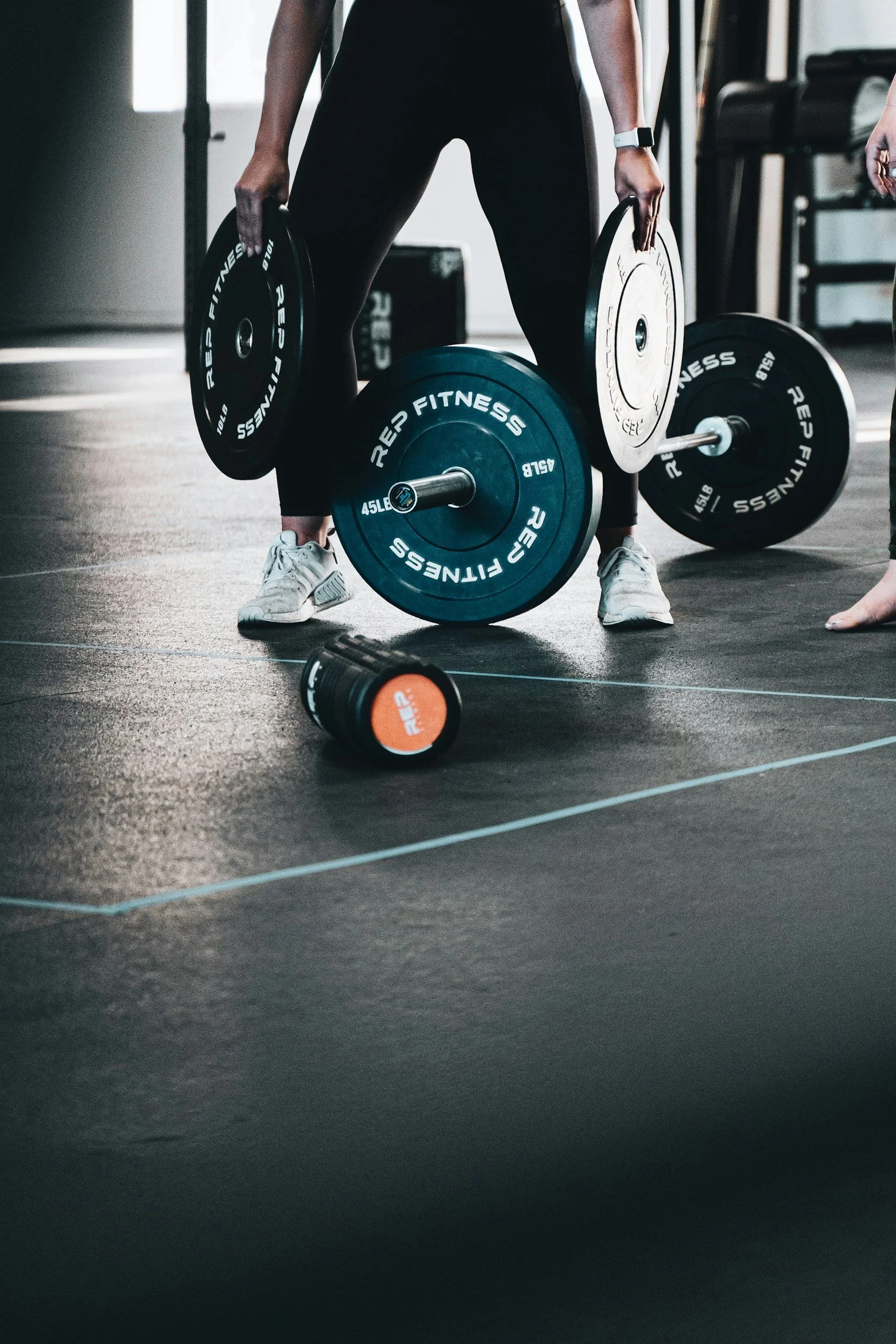 Person lifting weights in a gym, holding a barbell with weights, wearing athletic shoes and a watch, with a water bottle on the floor nearby.