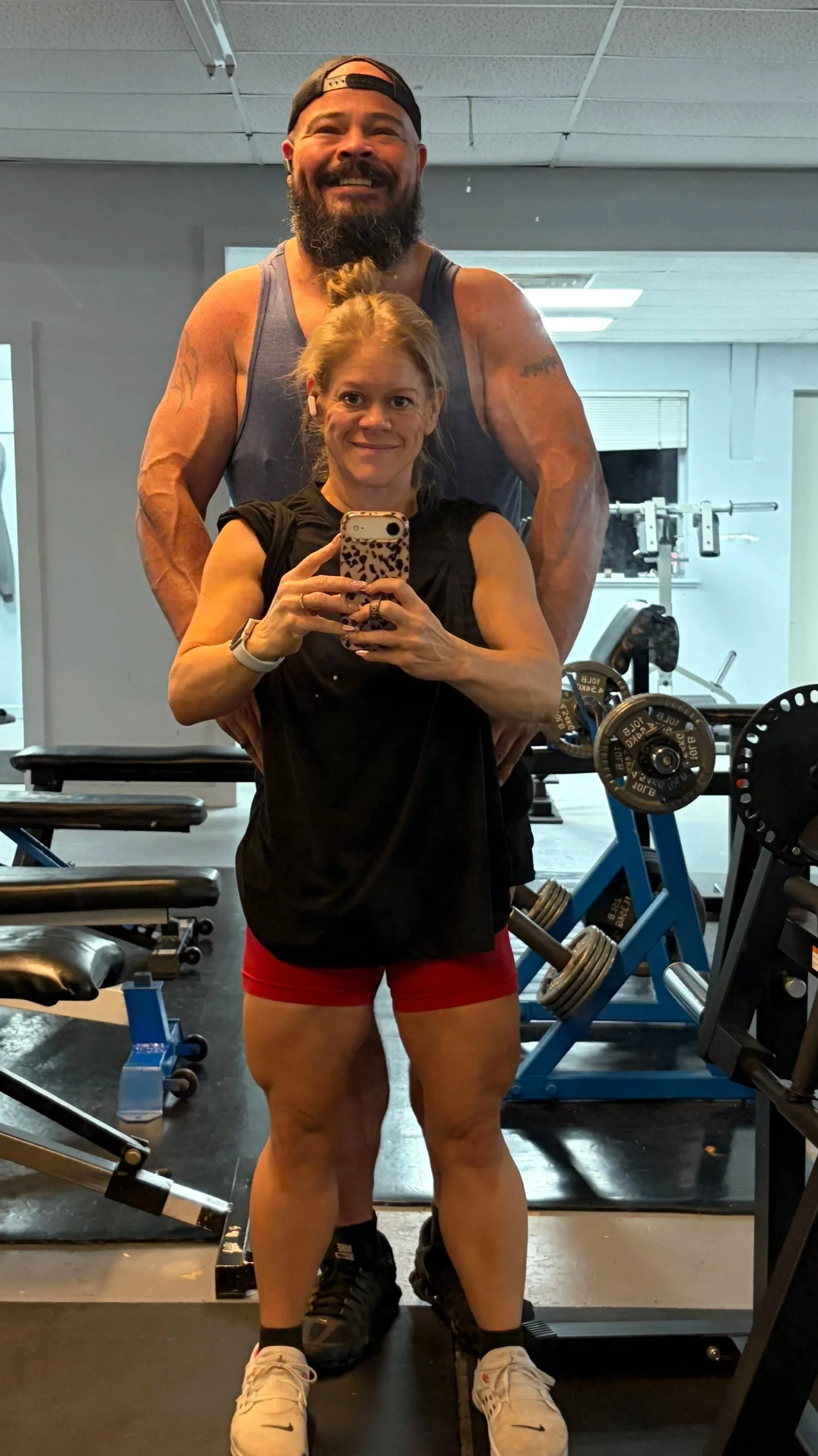 A woman taking a mirror selfie at the gym with a man behind her, both smiling and wearing workout clothes. The gym equipment includes weights and benches visible in the background.