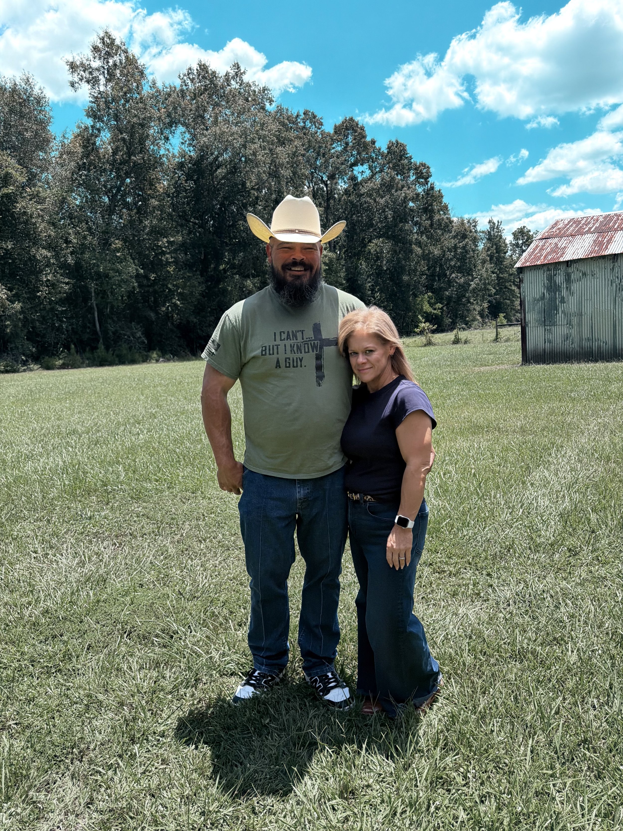 A man and woman standing outdoors in a grassy field with trees and a rustic shed in the background, under a partly cloudy sky. The man is wearing a cowboy hat and a gray T-shirt, and the woman is wearing a dark T-shirt and modern watch.