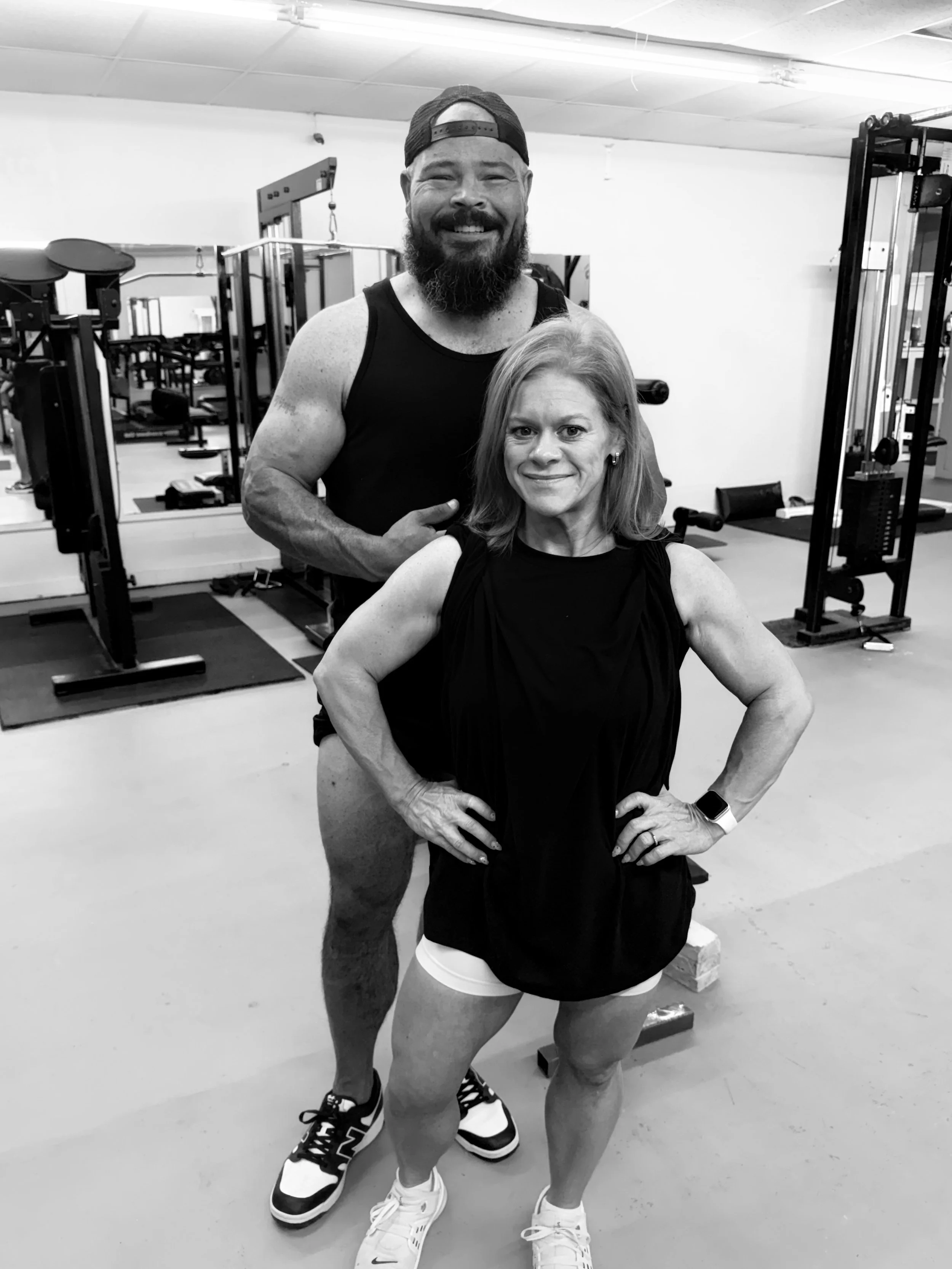 A black and white photo of a smiling man with a beard, wearing a backward baseball cap, sleeveless shirt, and athletic shorts, standing behind a seated woman with short hair, who is also smiling and wearing a sleeveless top and workout shorts, in a gym setting with exercise equipment in the background.