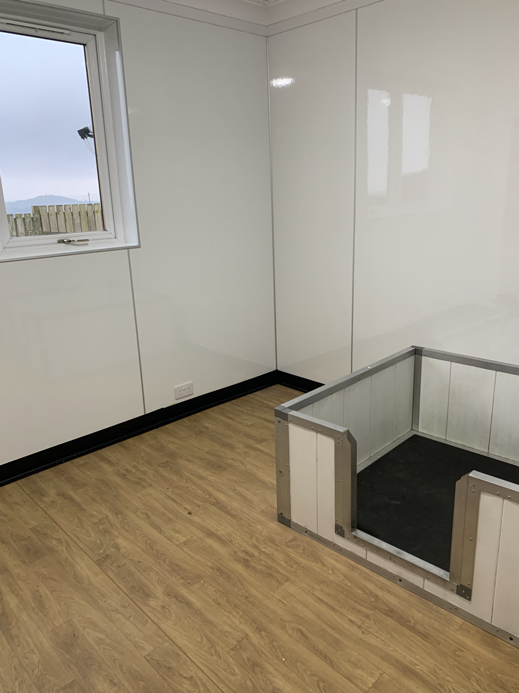 A corner of an empty room with wood-look flooring, white paneled walls, a small window with a view of a fence and mountains, and a dog crate with metal frame and black floor in the corner.