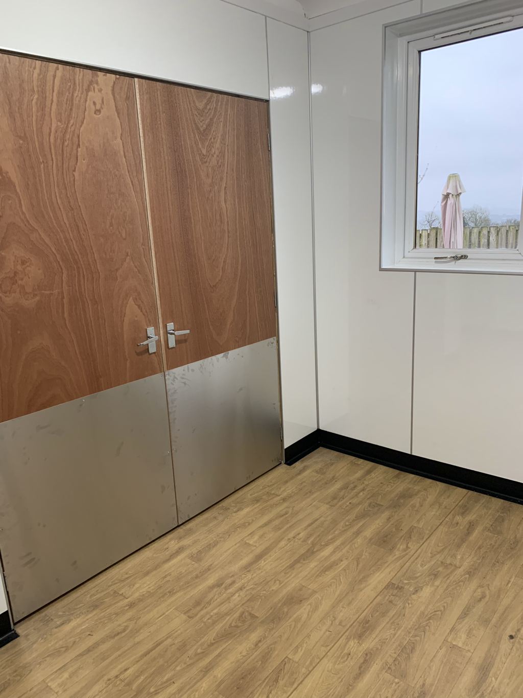 Empty room with a wooden cabinet, white walls, a window showing an outdoor patio with an umbrella, and wood-like flooring.