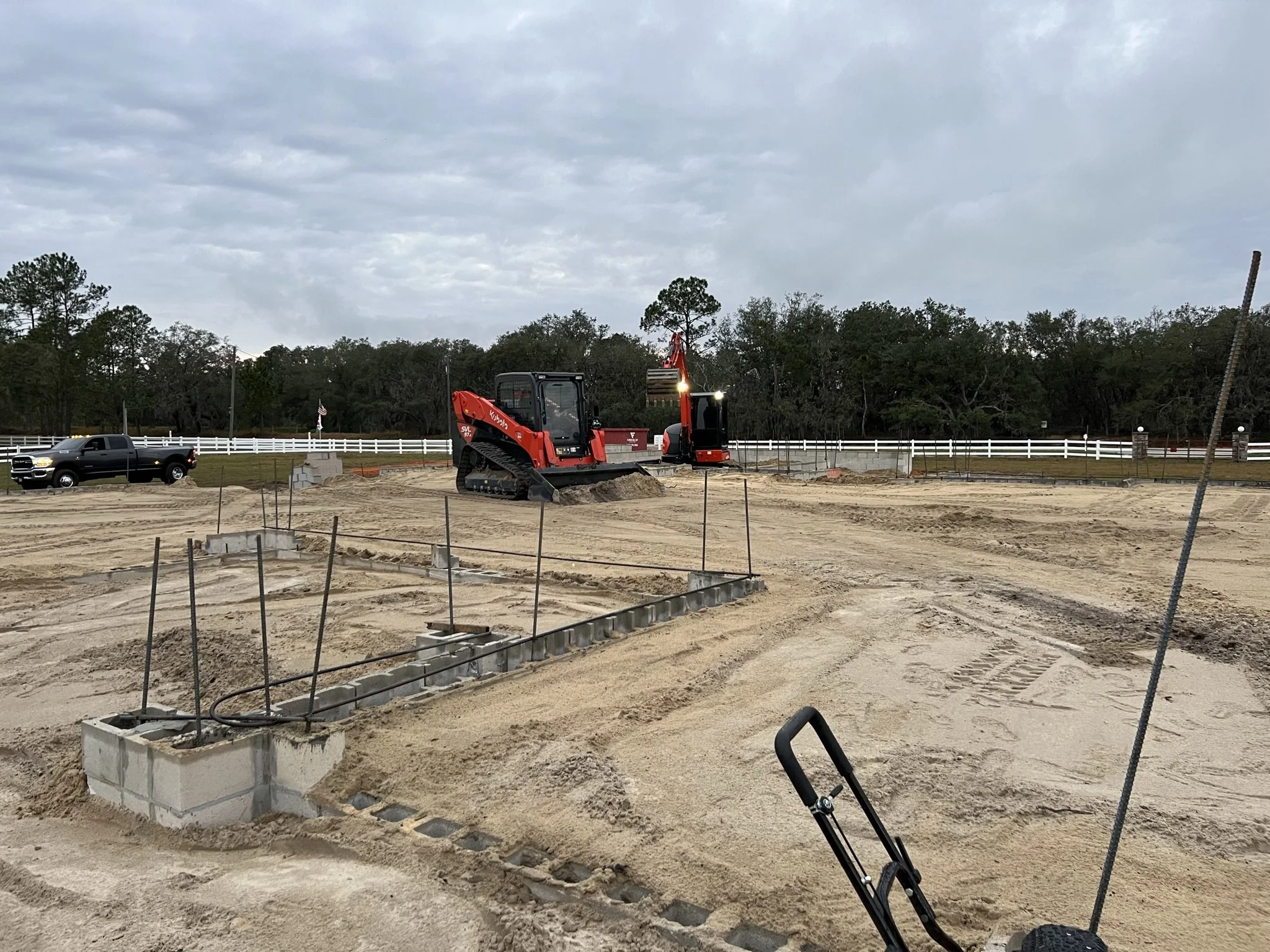Construction site with a red Kubota compact excavator, concrete foundation, and fencing under a cloudy sky.