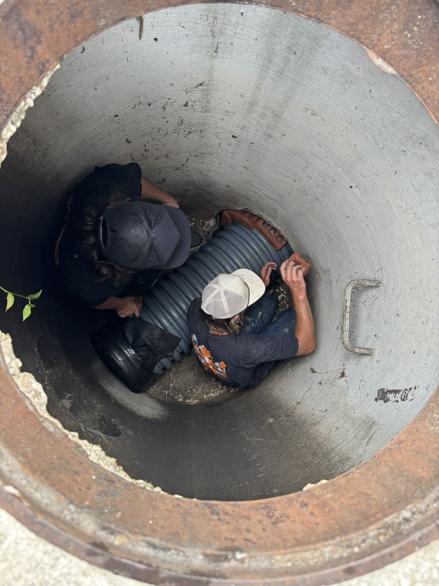 Two workers installing large corrugated pipes inside a concrete underground utility tunnel or manhole, seen from above.