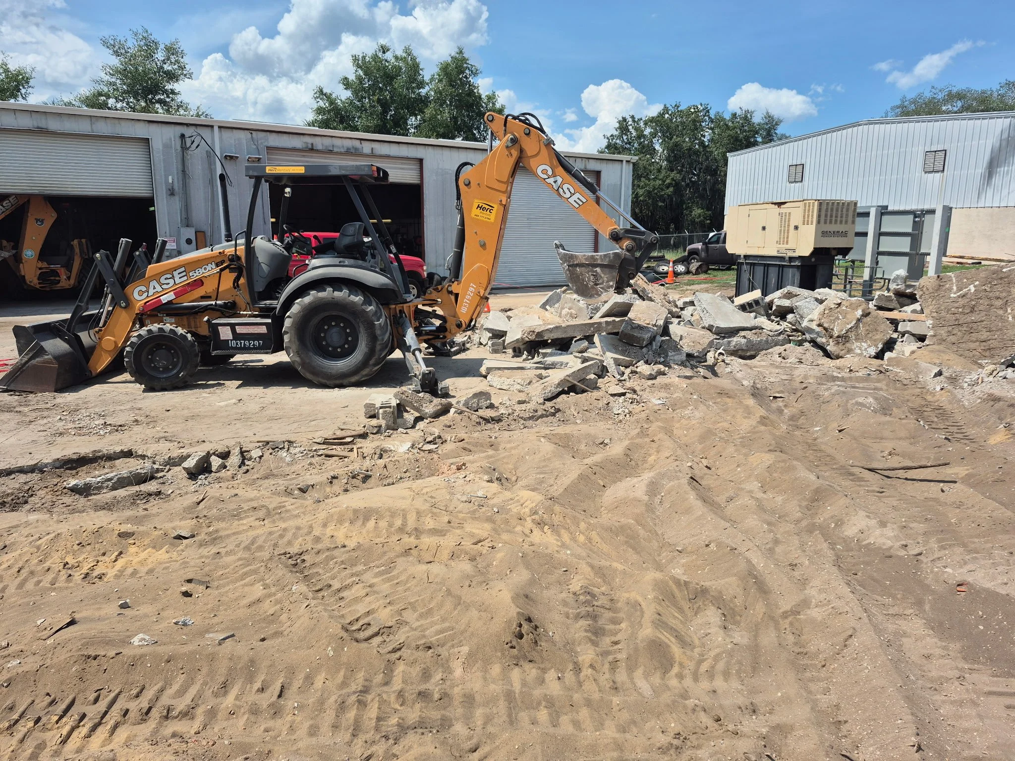 A construction site with a backhoe loader moving debris and rocks, surrounded by warehouses and construction equipment.